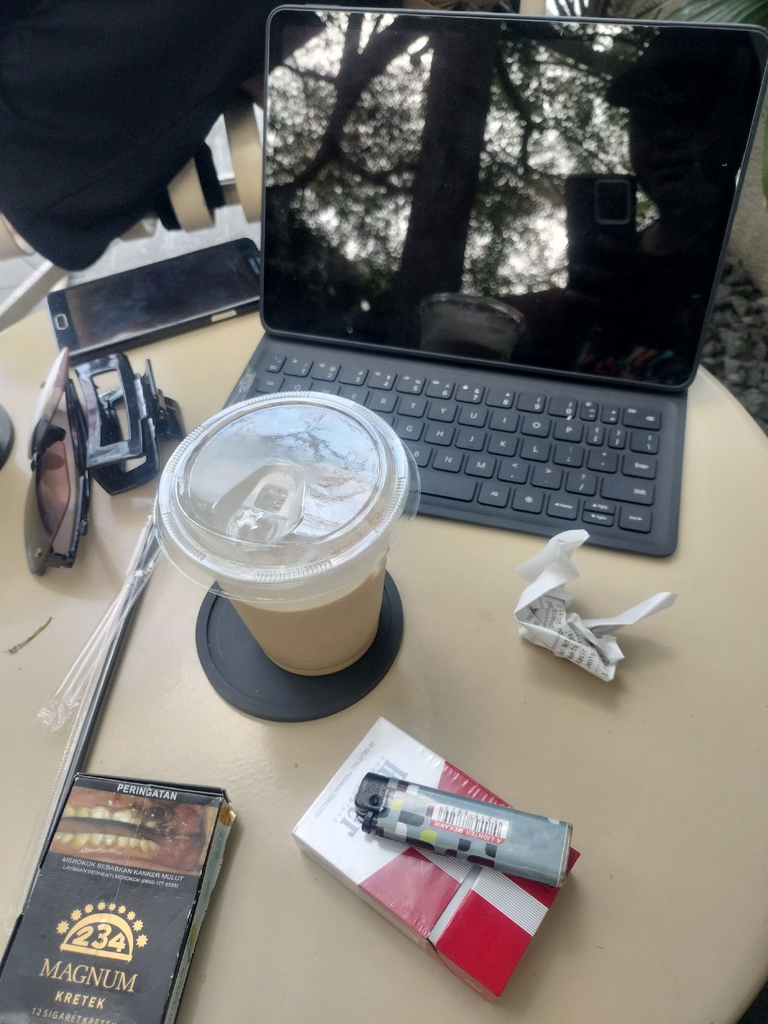 In Tamansari, Indonesia, on a beige table, a digital tablet with a black keyboard is positioned in the upper right quadrant. A disposable coffee cup with a clear plastic lid, containing a light brown beverage, sits on a black coaster to the left of the tablet. Adjacent to the coffee cup are a pair of dark-rimmed sunglasses and a black hair clip. In the lower left, a pack of "Magnum Kretek" cigarettes is visible, with a warning label depicting unhealthy teeth. To the right of the cigarette pack, a silver lighter rests on top of a red and white cigarette pack. A crumpled piece of paper with visible text is situated between the coffee cup and the cigarette packs. A smartphone lies flat on the table between the tablet and the sunglasses. A black garment is draped over the left side of the image. The reflection on the tablet screen shows trees and foliage, suggesting an outdoor setting.