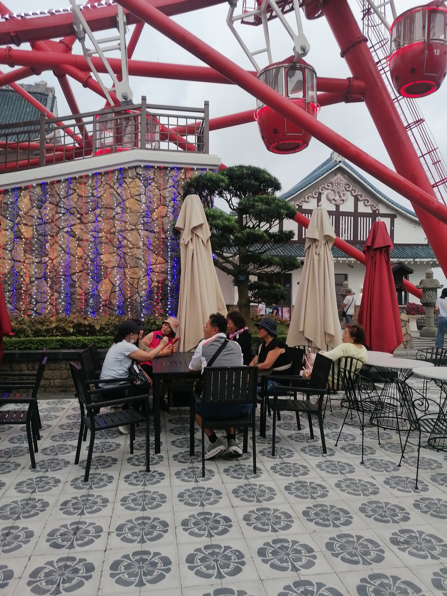 The scene depicts an outdoor café setting in Hong Gai Ward, Vietnam, situated at the base of a large red Ferris wheel. The cafe area is paved with grey and white tiles in a floral pattern, with black metal tables and chairs arranged for several groups of people.
A group of individuals are seated at one of the tables, seemingly engaged in conversation. Other individuals are seated at nearby tables, some facing away from the camera. A few unopened beige umbrellas and a red umbrella stand near the tables.
In the background, a stone wall adorned with strings of small red, white, and blue lights provides a backdrop. A traditionally styled white and brown building with a dark tiled roof is visible behind the wall. The red Ferris wheel dominates the scene, with its cars numbered "18". The sky appears overcast, suggesting a cloudy day.