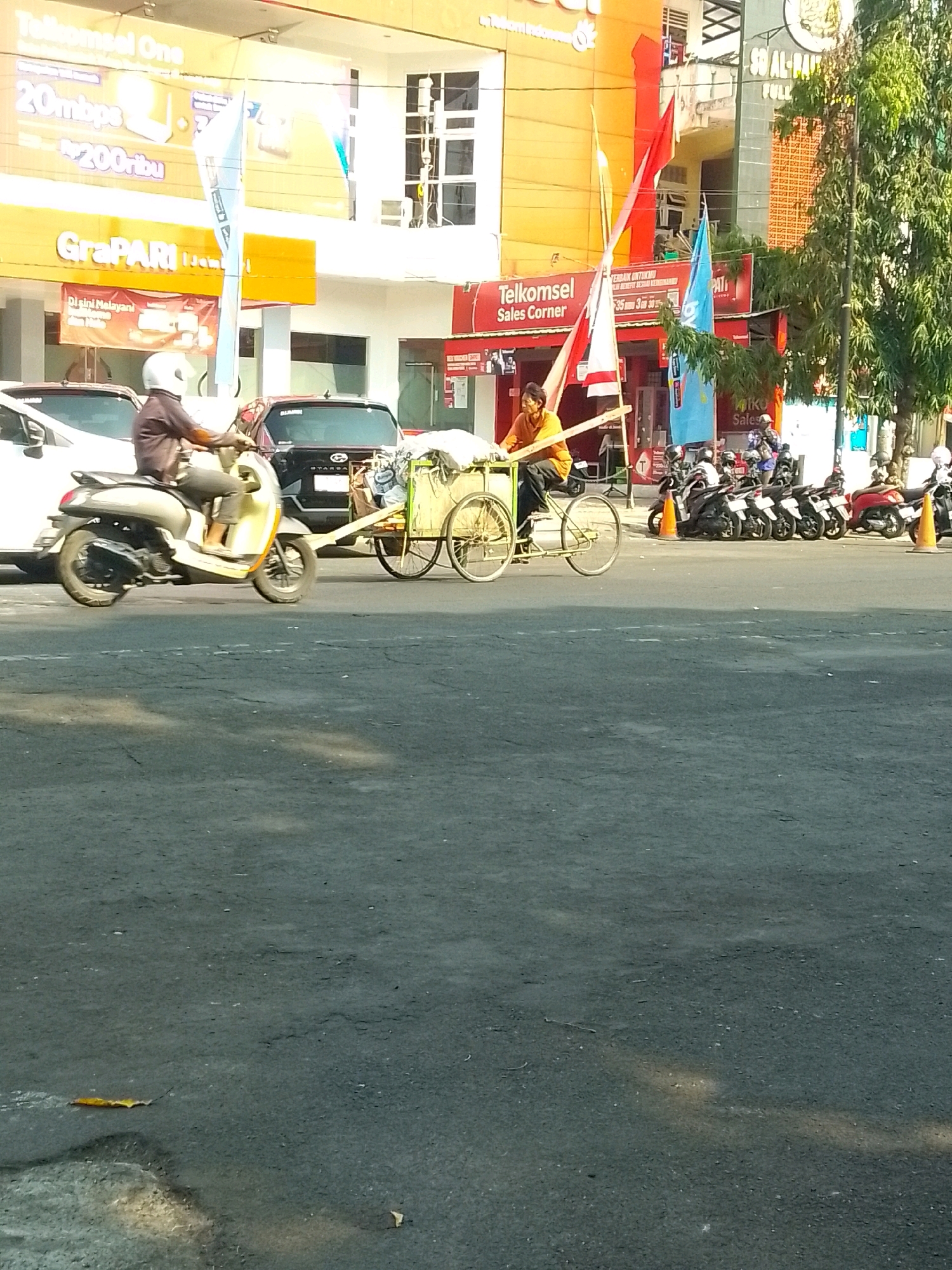 The image depicts a street scene in Jember, Indonesia. In the foreground, a man wearing an orange shirt and dark pants is seated on a tricycle-like cart, pulling it with his legs. The cart is loaded with what appears to be sacks and other goods. To the left of the cart, a person is riding a scooter, also in motion. Behind the scooter, there are two cars. The background features several buildings, including one with a large yellow sign that reads "GraPARI" and "Telkomsel One." Another building has a "Telkomsel Sales Corner" sign. Multiple motorcycles are parked in front of the buildings, some with riders. The overall environment suggests a commercial area with active street traffic.
