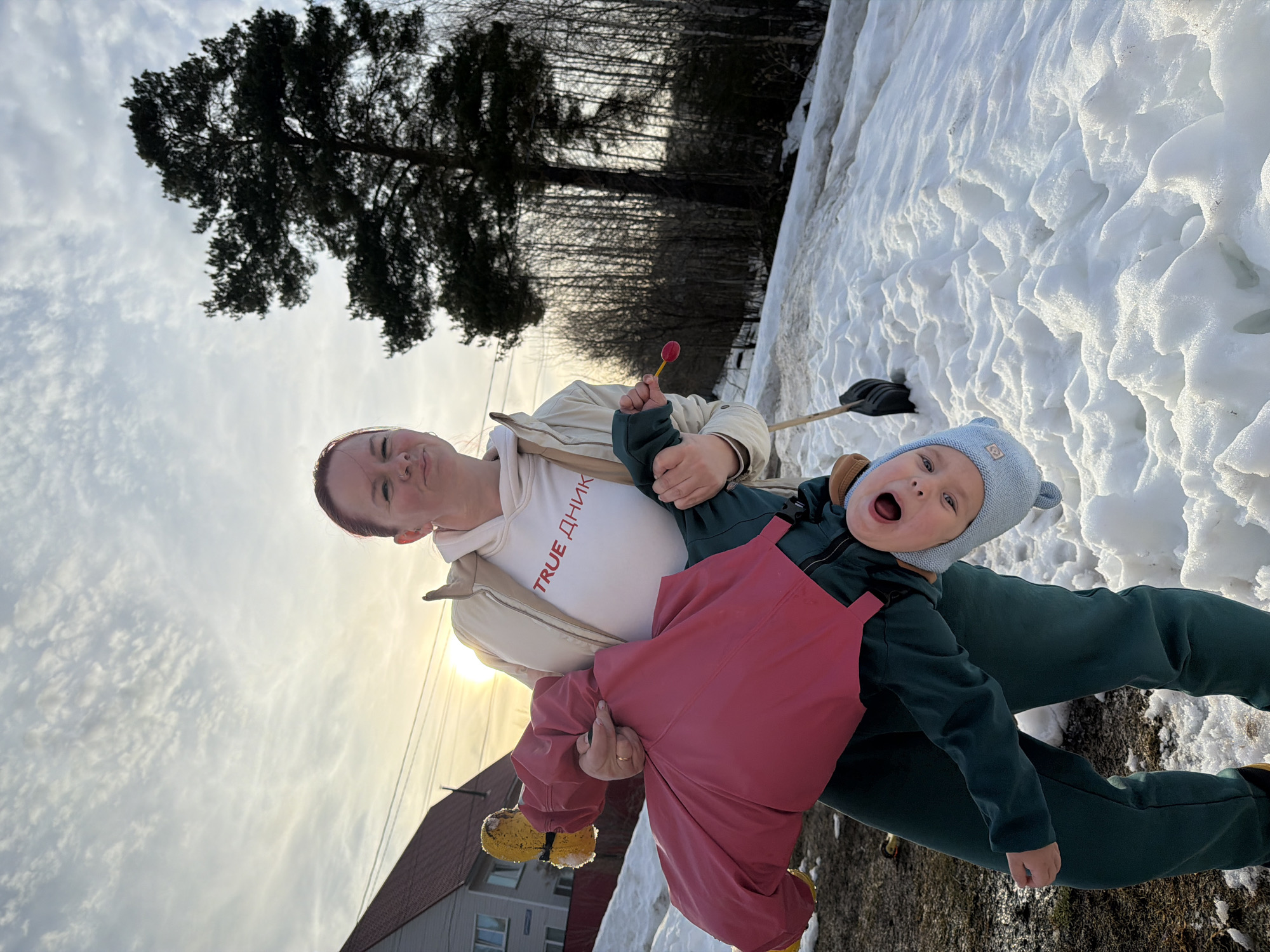 An outdoor scene in Chaikovsky, Russia, depicts an adult female holding a young child upside down on a snow-covered slope. The adult, with reddish-brown hair tied back and a left nostril piercing, wears a beige jacket over a white hooded sweatshirt inscribed with "TRUE ДНИК" in red lettering, and dark green pants. A gold-colored band is visible on her left ring finger. In her right hand, she holds a small, red, lollipop-shaped object with a yellow stick. The child, dressed in a light blue knitted hat with ear-like protrusions, a dark green outfit, and a pink/red waterproof overall, has yellow boots partially covered in snow and an open mouth, looking towards the viewer while suspended upside down. The ground is characterized by uneven patches of melting snow and exposed brown earth. In the midground, a black shovel with a wooden handle is partially visible, leaning against the snow. The background features a light-colored building with a red sloped roof and multiple windows on the left, with utility lines crossing in front of it. To the right, a large dark green conifer tree stands prominently before a dense grove of bare deciduous trees. The sky above is light blue with scattered white clouds, and the sun is low on the horizon, casting a warm glow.