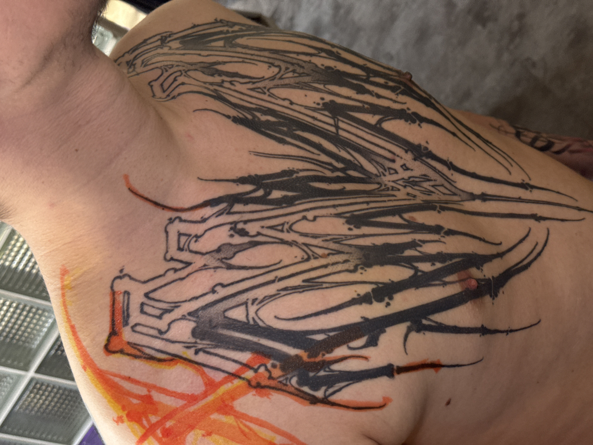 The image shows a close-up of a person's upper chest and shoulder with a large, abstract tattoo in black and orange ink. The tattoo covers the area from the upper chest to the shoulder. The tattoo appears to be an unfinished piece. It consists of black lines and patterns. Additional orange ink details are noticeable on the lower part of the tattoo. The image does not contain enough information to pinpoint a specific location in Moscow, Russia, as no identifying landmarks or contextual clues are present.