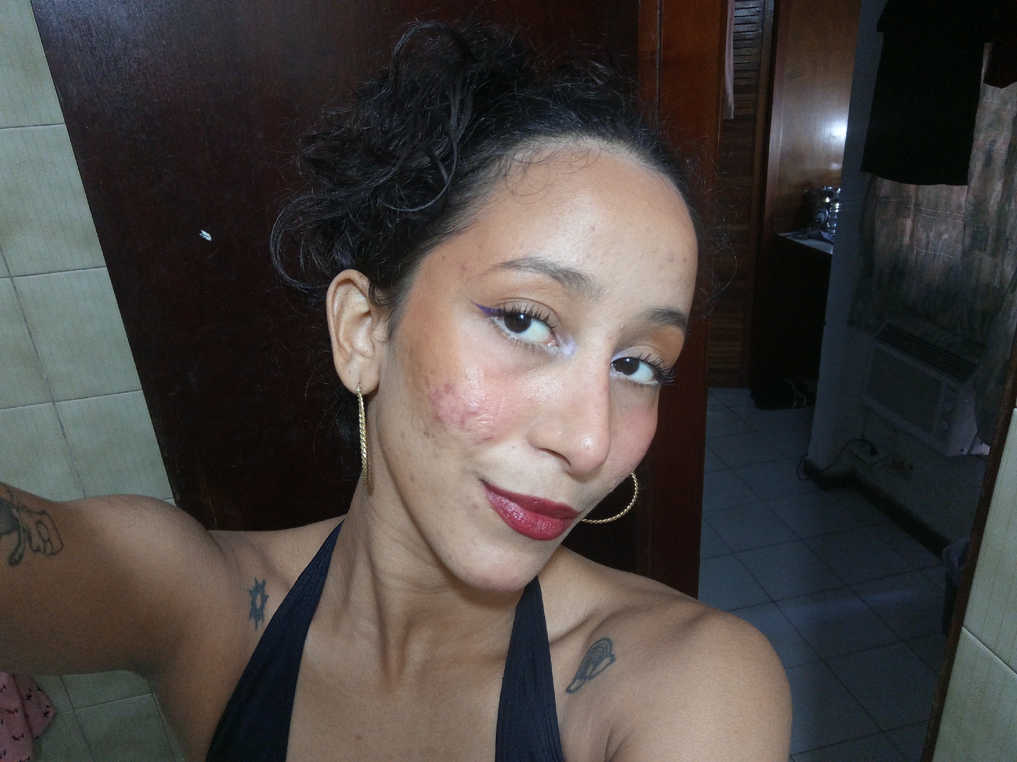 A selfie portrait of a light-skinned woman with dark curly hair pulled back, wearing a black halter top. She has noticeable acne scars on her left cheek and is wearing dark red lipstick and purple eyeliner. Her eyes are focused on the camera. She is wearing large gold hoop earrings. Tattoos are visible on her left arm and shoulder. The background is a dark wooden door and a glimpse into another room with tiled floors and a dark piece of furniture. The location is not discernible from the image.