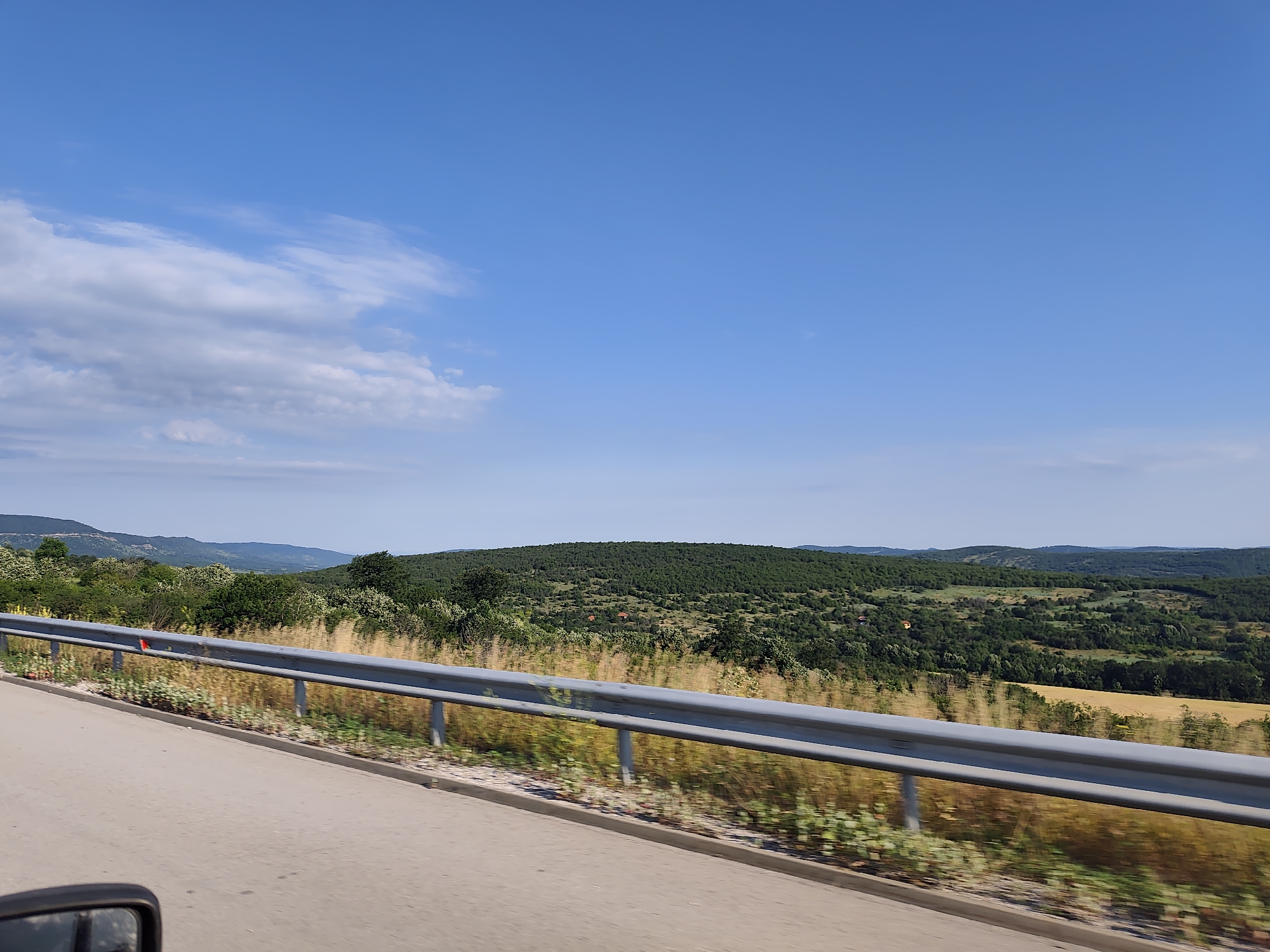 This is a photograph taken from a moving vehicle, likely a car, showing a landscape view from the side of a road. The foreground shows a portion of the road and a metal guardrail. The guardrail is silver and runs horizontally across the image, separating the road from the natural environment beyond. The road itself appears to be paved asphalt, with a faint white line visible on the left side.
Beyond the guardrail, the landscape is characterized by rolling hills covered in dense green trees. In some areas, the trees are interspersed with patches of dry, golden grass. Further in the distance, the hills become less distinct and fade into a hazy, blue sky with scattered white clouds. There are a few small, red-roofed buildings visible nestled within the greenery on the hills. The overall impression is a rural, somewhat undeveloped area. Given the provided context, the image depicts a scene near Yablanitsa, Bulgaria.