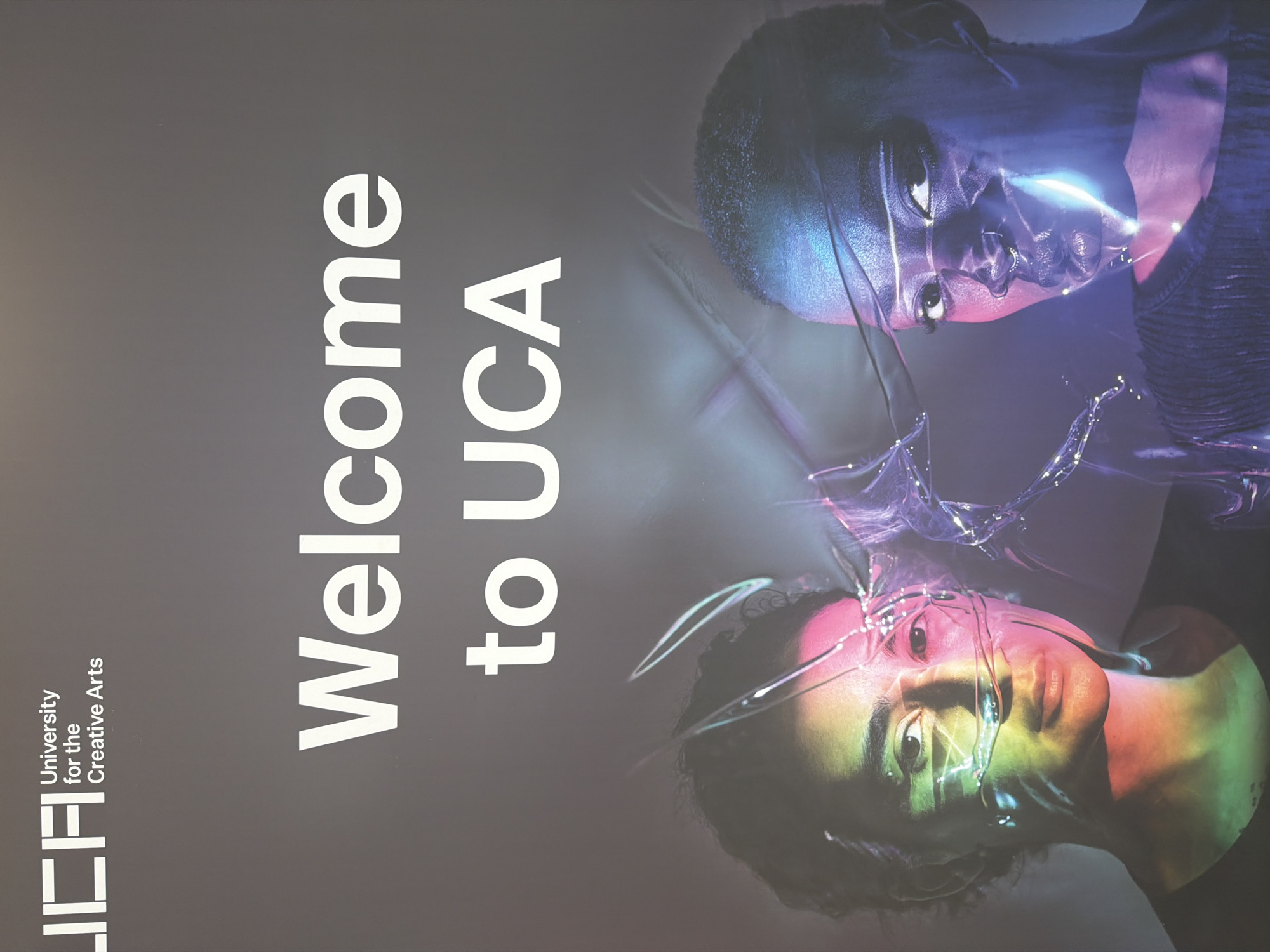 The image shows a poster with text and two portraits. The text at the top left reads "UCA" in a stylized font, with "University for the Creative Arts" written below it. Below this, larger white text states "Welcome to UCA". The bottom half of the poster features two portraits of individuals with artistic lighting and effects. The person on the left has colorful lights cast on their face, with what appears to be a transparent, fragmented material overlaying their features. The person on the right also has dramatic lighting in shades of blue and purple, with similar fragmented material over their face, highlighting facial features. The background is a dark, textured gray. The poster is likely displayed at the University for the Creative Arts campus in Epsom and Ewell, United Kingdom.