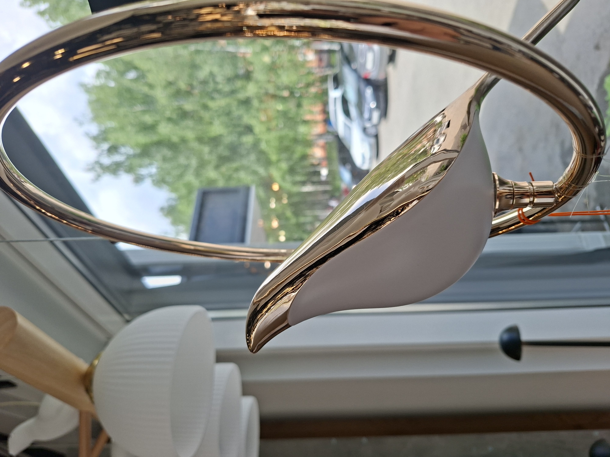 A close-up, eye-level shot focuses on a decorative, metallic gold bird sculpture attached to a large, circular gold frame. The bird, with its glossy, reflective surface, is positioned in the foreground, its head turned to the left. Behind the sculpture, a window reveals an outdoor scene with trees and parked cars, suggesting an urban environment. In the background, on the left, a white, pleated lampshade is partially visible, hinting at an interior setting like a shop or showroom. The overall lighting is bright, with natural light streaming through the window. The scene is located within a commercial space in Izhevsk, Russia, likely a store that sells lighting fixtures or home decor.