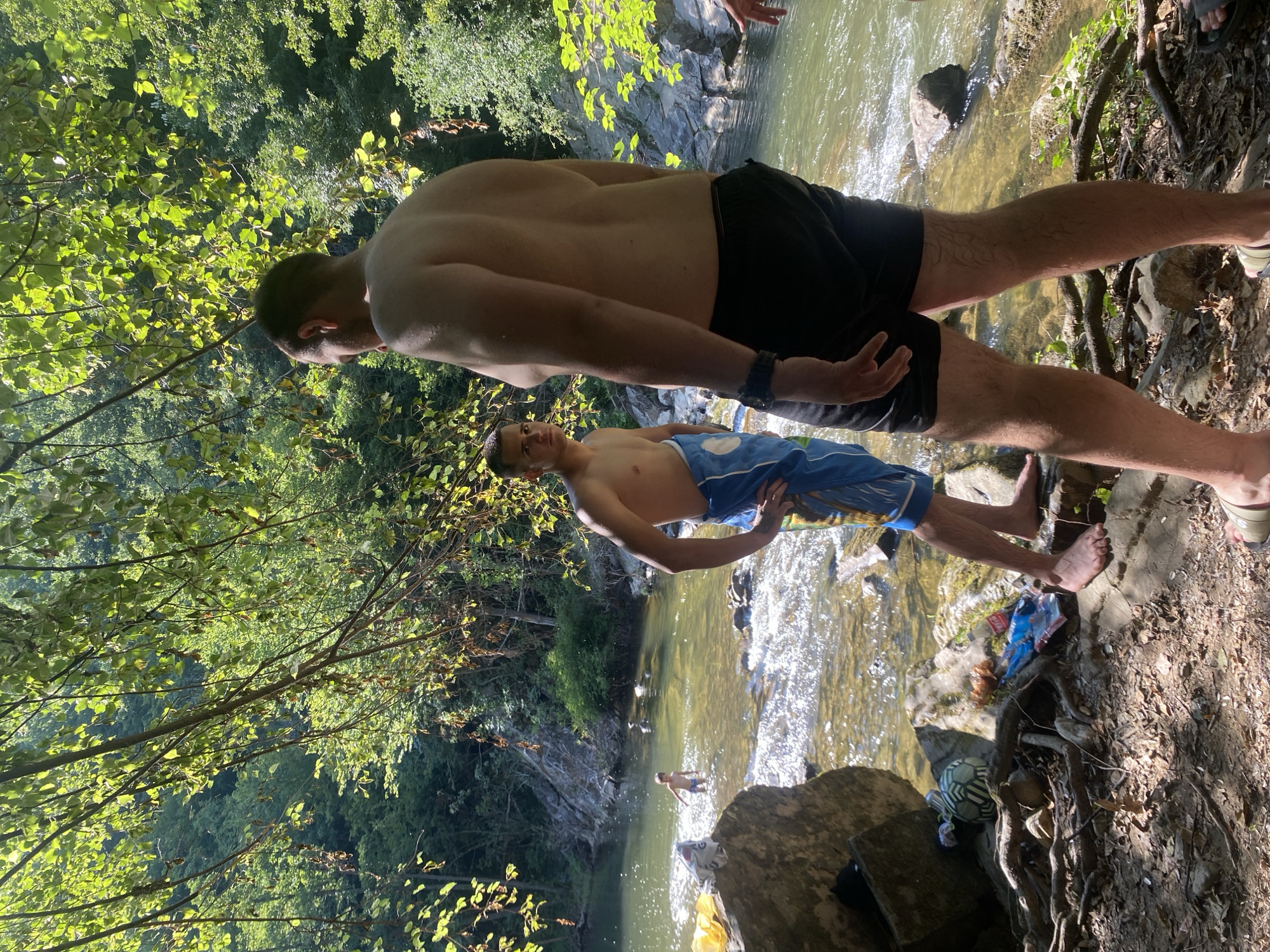 In Debnevo, Bulgaria, two young men are by a shallow, rocky river. The man in the foreground, facing away from the camera, wears black shorts and a towel wrapped around his waist. To his left, another young man, facing the camera, wears blue patterned swimming shorts and a towel draped over his shoulder. Further in the river, a younger child stands in the water. The surrounding environment is lush and green, with trees and foliage overhead casting dappled sunlight onto the scene. Rocks line the riverbed and banks. On the shore, near the men, are several items including a soccer ball, plastic bags, and a beverage can.