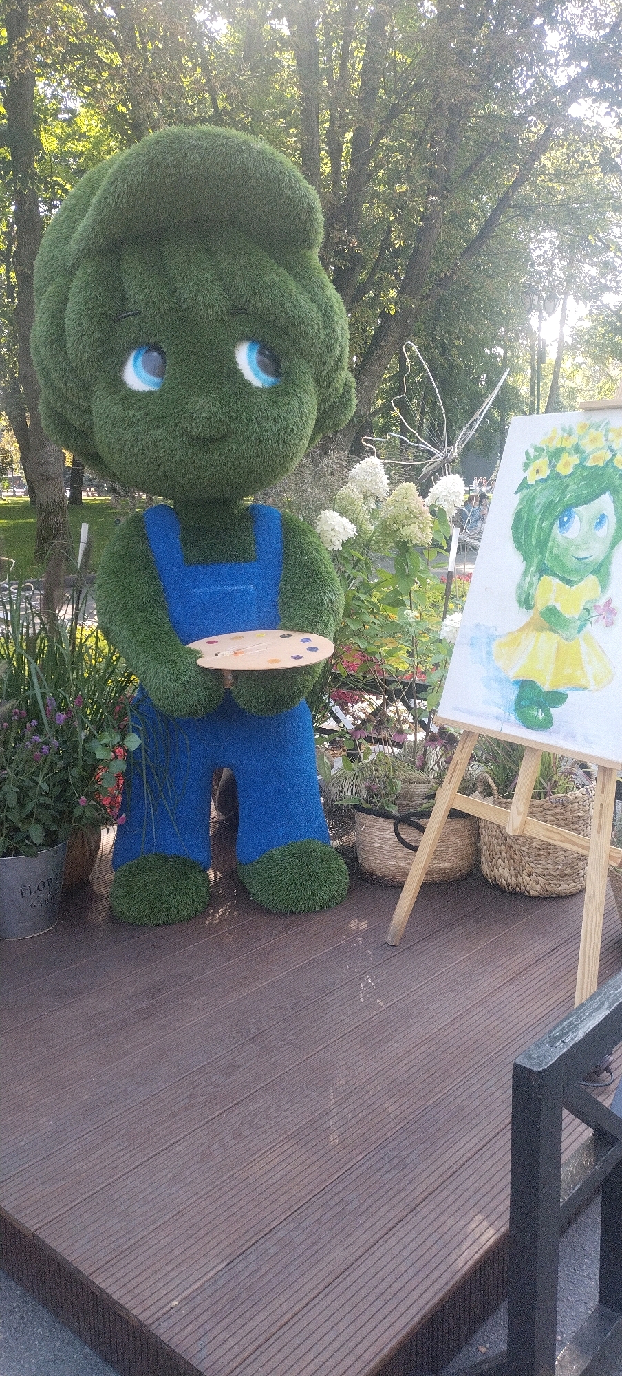 In a city park in Kharkiv, Ukraine, a topiary figure resembling a child, made of green grass-like material, stands dressed in blue overalls. The figure holds a wooden artist's palette with various colors of paint. To the right of the topiary, an easel displays a painting of a green-haired child wearing a yellow dress and a flower crown, holding a pink flower. The scene is set outdoors, with trees and foliage in the background. The ground is a wooden deck, and several potted plants and wicker baskets are visible around the figures. The overall impression is an art installation in a park setting.
