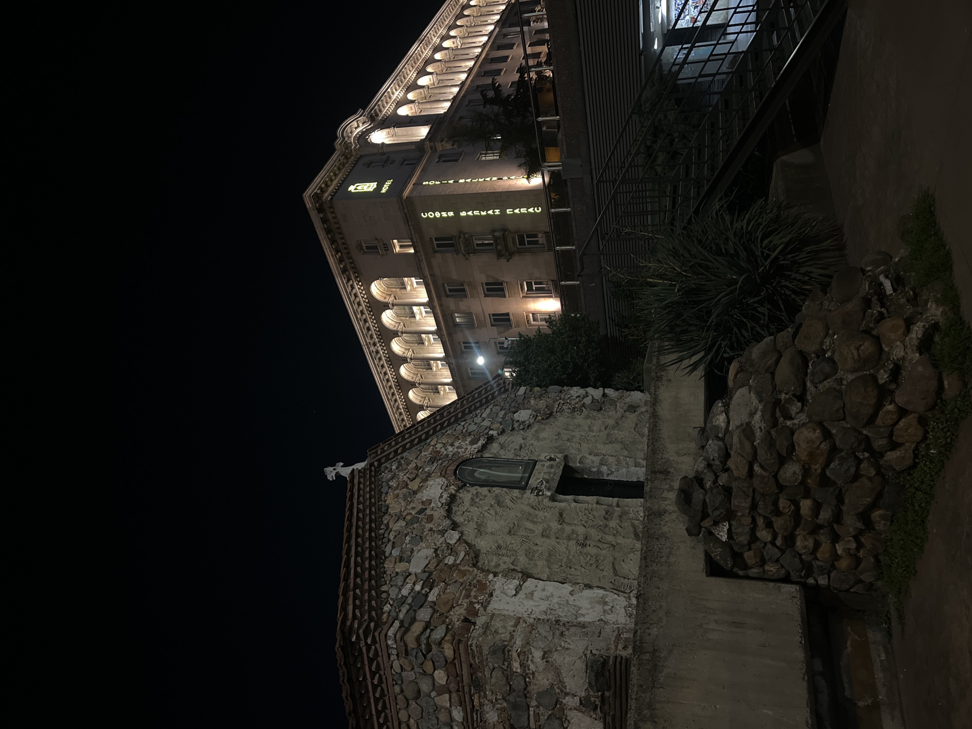An outdoor, low-angle, nighttime photograph captures a historic urban scene in Sofia, Bulgaria, characterized by two distinct architectural styles. The dominant feature is a grand, multi-story building on the left, illuminated by warm artificial lights that highlight its classical facade with multiple arched windows and ornate cornices. This building appears to be a hotel, as indicated by the glowing green text "SOFIA BALKAN PALACE" and "HOTEL" displayed vertically on its side, along with a discernible logo.
In the foreground and to the right, a smaller, ancient-looking stone structure stands with a roughly textured wall composed of various sizes of rocks and some brickwork. This structure features a small, arched window and a white, cross-like finial or sculpture on its peak, suggesting historical or religious significance.
The sky is completely black, indicating a clear night. Lush green vegetation, including a large spiky plant, is visible around the base of both structures and along a concrete or paved area. A metal staircase or railing is partially visible on the far right, leading upwards next to the larger building. No people or activities are visible, lending a serene and timeless quality to the scene. The lighting emphasizes the architectural details and the contrast between the well-maintained hotel and the weathered stone structure.