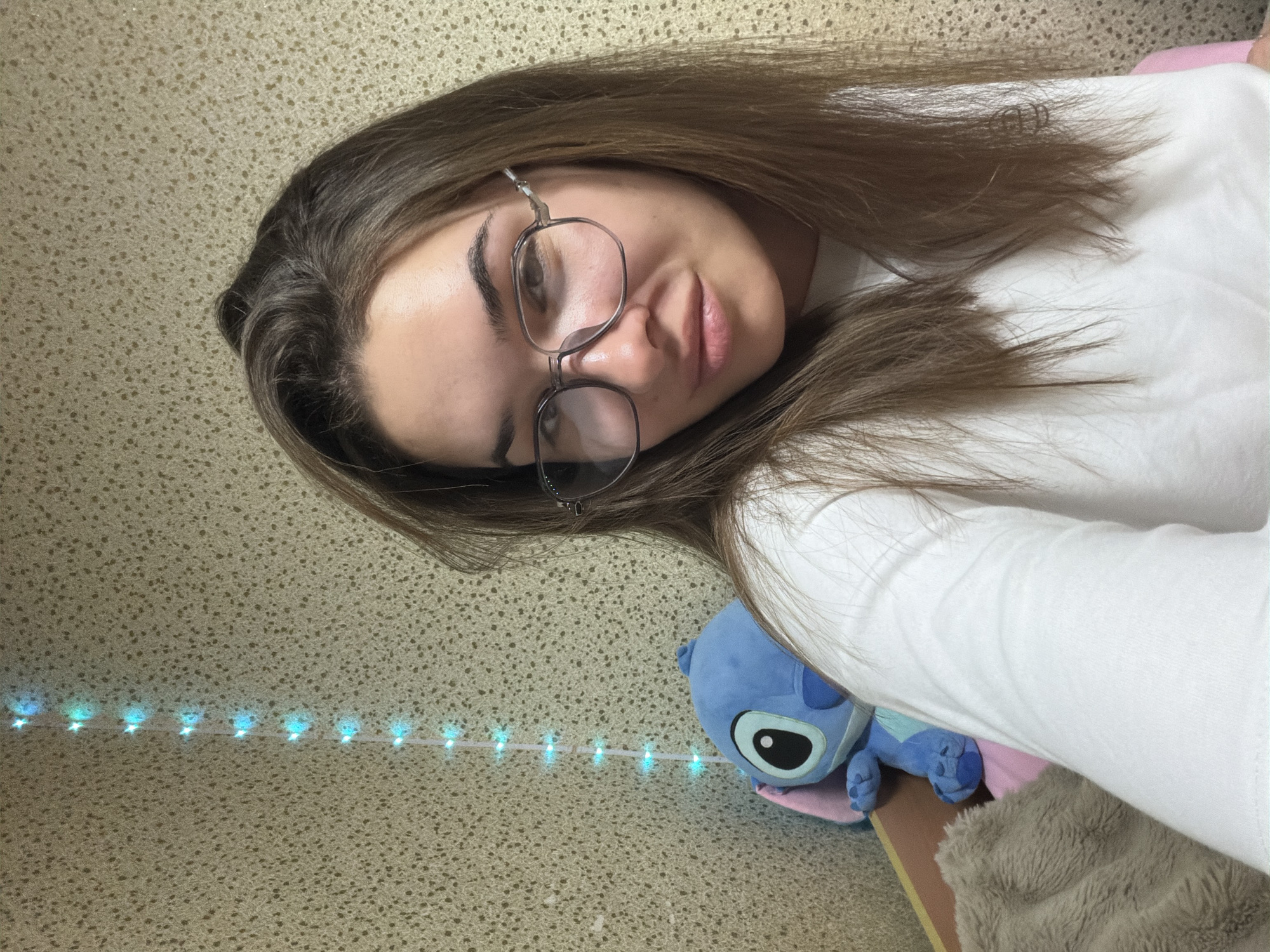 A woman is shown in a selfie, wearing glasses and a white shirt. Her hair is brown and styled loosely around her face. To her right is a stuffed blue character toy, likely Stitch from Disney's Lilo & Stitch, resting on a pink surface. A string of blue lights is visible on the textured beige wall behind her. There is no indication of a location in Sofia, Bulgaria, or any other city.