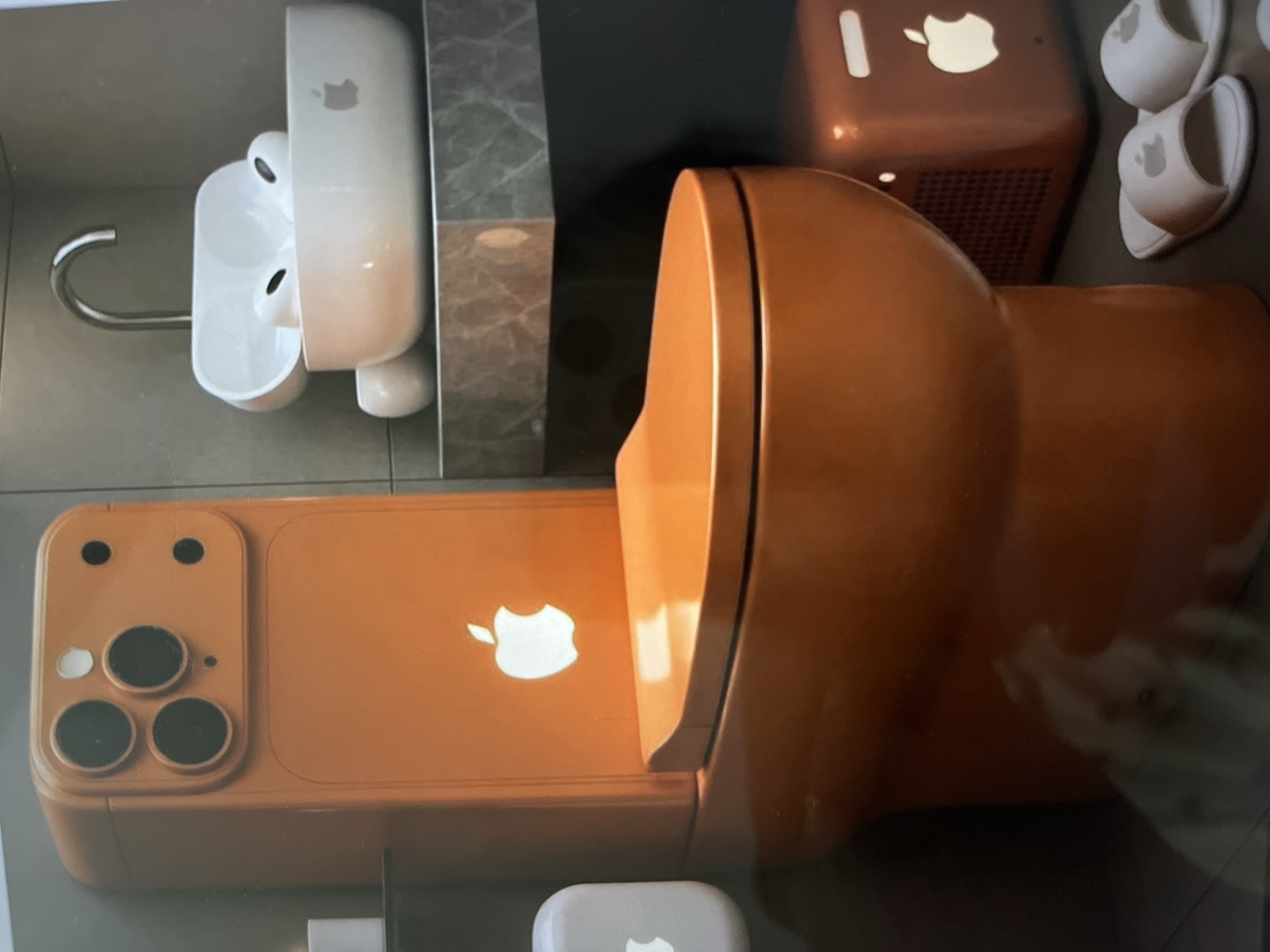 An orange toilet with an Apple logo on the cistern is shown in the center of the image. The cistern is designed to resemble the back of an iPhone, complete with camera lenses. To the right of the toilet, there is a white sink shaped like an AirPods charging case, with a silver faucet. Below the sink, a marble shelf is visible. To the right of the toilet, a small orange square object with an Apple logo, resembling a speaker, is also present. In the foreground, a pair of white slippers with Apple logos are on the floor. The overall setting appears to be a bathroom.