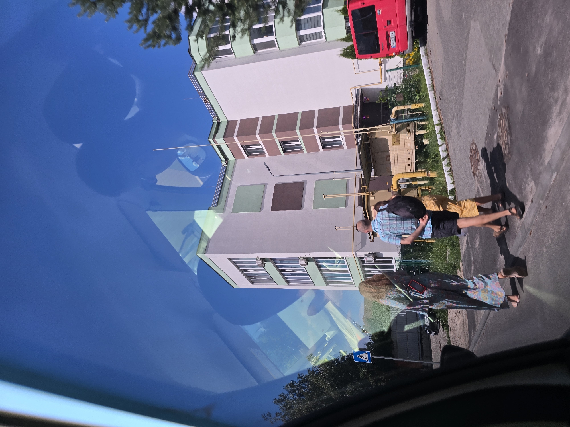 This media file depicts a street scene in Petropavlivska Borshchahivka, Ukraine, as seen through a car windshield, implying a reflection. The image is oriented sideways.
The main subjects are two people walking on a paved sidewalk next to a building. One person, wearing a plaid shirt and dark shorts, carries a backpack. The other person is wearing a colorful, long dress. They appear to be walking in the same direction. Shadows suggest it is daytime, likely afternoon, under clear, sunny weather.
The setting includes a modern-looking building with green and brown accents and numerous windows. Yellow pipes are visible on the side of the building, suggesting some kind of utility infrastructure. A red bus is parked to the right of the building. The ground is asphalt, and there is some greenery and trees in the background and along the sidewalk. A blue pedestrian crossing sign with a white pictogram is also visible on the sidewalk.
The reflections on the windshield create a sense of depth and overlay the scene with the blue sky and some tree foliage. The overall impression is a snapshot of everyday life on a sunny day in a Ukrainian town. No discernible text is visible in the image.