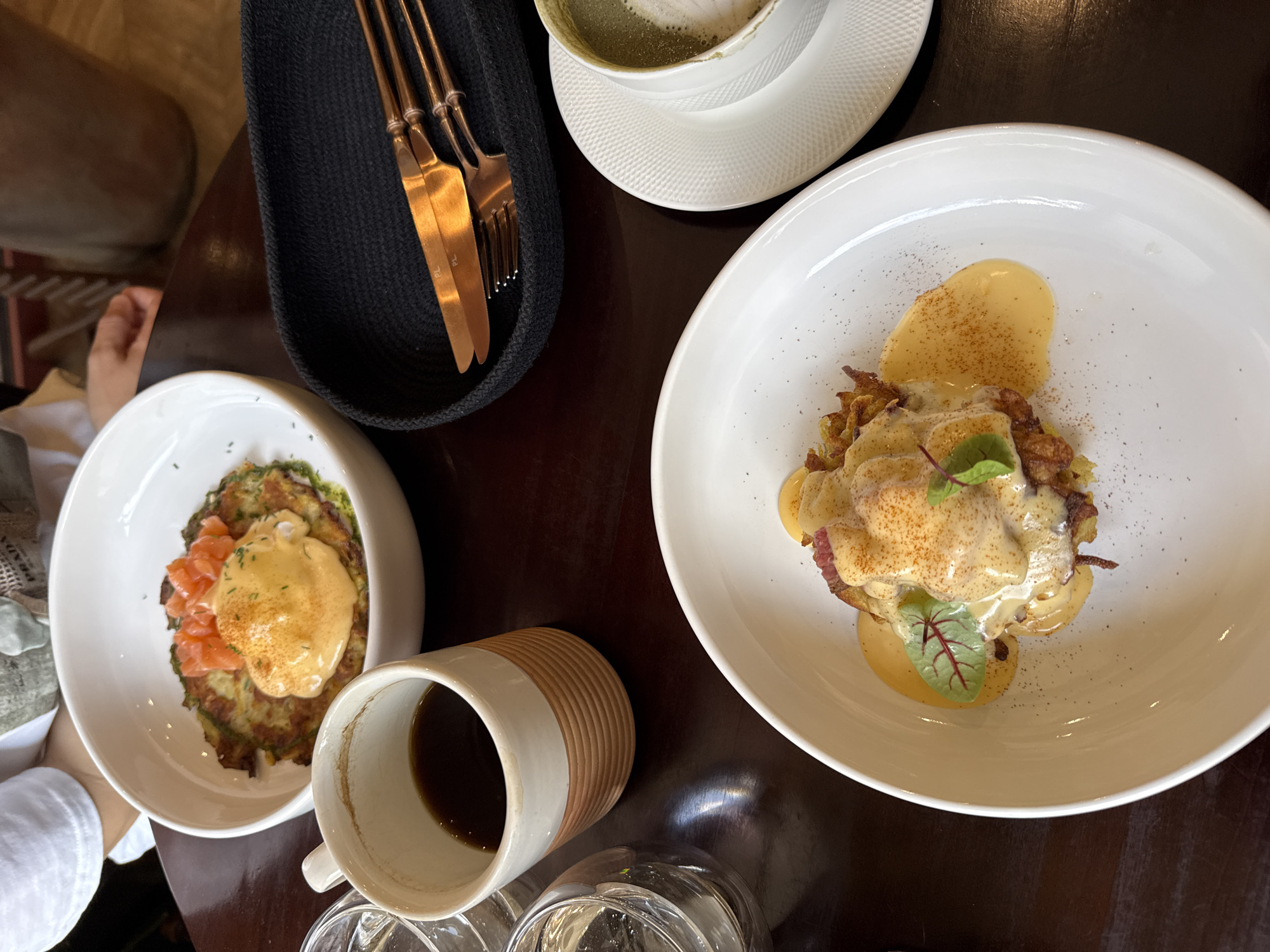The image shows a table setting with two plates of food, possibly potato pancakes or latkes topped with eggs benedict and different toppings. The plate on the left has what appears to be salmon as a topping, while the plate on the right appears to have red meat. There are also drinks: matcha latte in a cup on a saucer and a cup of coffee. Cutlery, including forks and knives, is placed in a black holder. There are also two empty water glasses at the bottom. The location is potentially Krasnoyarsk, Russia.