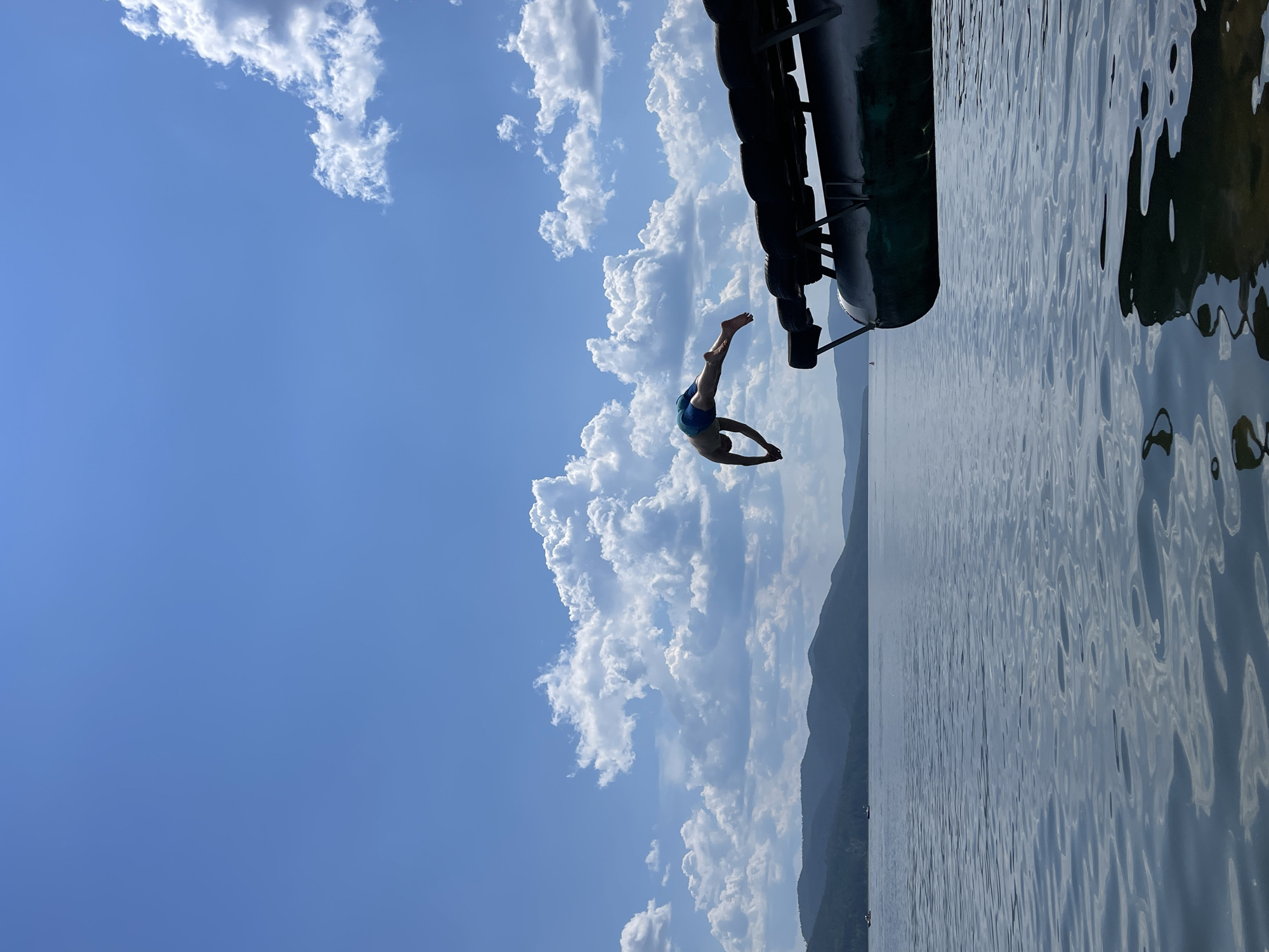 The image depicts a person diving into a body of water, likely Krasnoyarskoye Reservoir near Divnogorsk, Russia. The sky is blue with scattered white clouds. In the foreground, the edge of a pontoon boat or dock is visible, adorned with black tires for protection. A person, wearing blue swim trunks, is captured mid-air, executing a dive with their body arched and arms extended. The water is calm with ripples reflecting the sky and surrounding landscape. In the distance, rolling green hills or mountains form the horizon. Two small boats are visible on the water, adding to the sense of scale. The overall scene captures a moment of recreation on a sunny day.