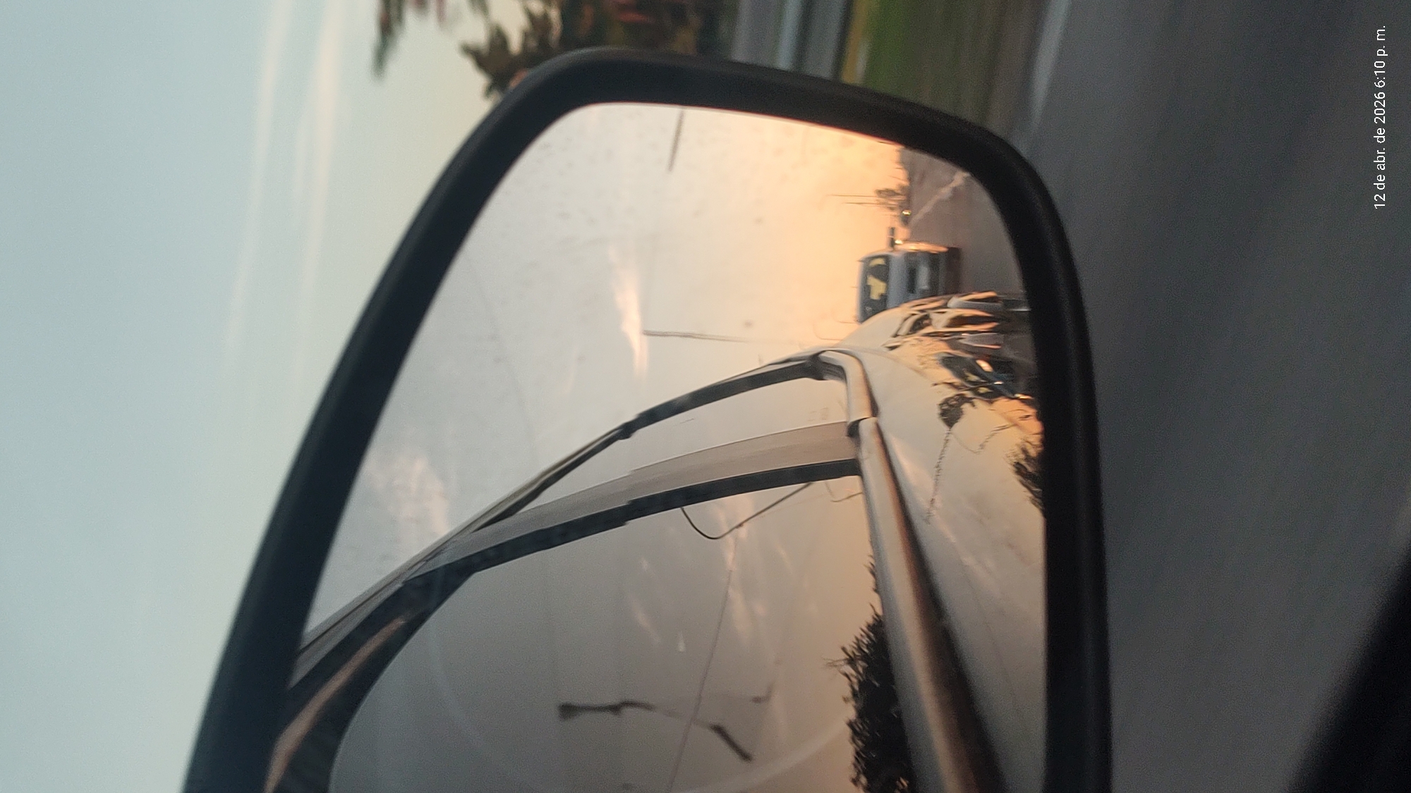 The image shows a car's side mirror reflecting a road and a sunset. Another car is visible in the distance on the road. The text "12 de abr. de 2026 6:10 p. m." is in the upper right corner. The location might be Villa Domínico, Argentina.