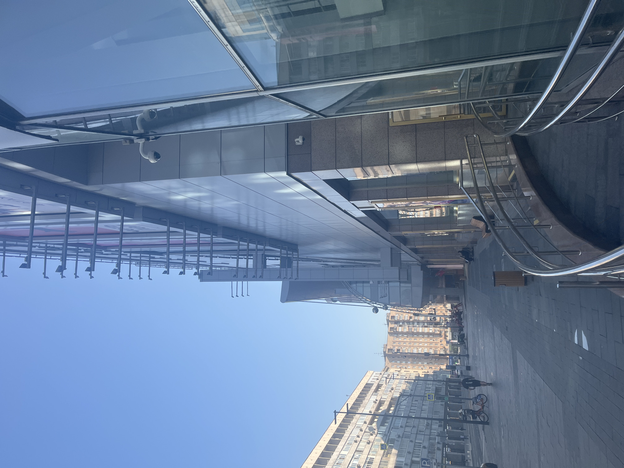 The image depicts a street scene in Moscow, Russia, on a clear day. In the foreground, a modern building with large glass windows and a curved metal railing leads the eye into the scene. The building's facade is a mix of glass and metallic panels, with some advertising visible through the glass. To the left, a wide paved sidewalk runs alongside the building, leading into a street lined with apartment buildings.
Several people are present on the sidewalk and street. A person is walking down the street away from the camera, wearing dark shorts and a t-shirt. Another person is near a bicycle, which is orange and black. Further down the street, two people are seated on a bench. The street is illuminated by sunlight, casting shadows on the pavement. The sky is a clear, bright blue. The apartment buildings on the left are characteristic of Soviet-era architecture, with numerous windows and balconies. The overall impression is of an urban environment during daylight hours.