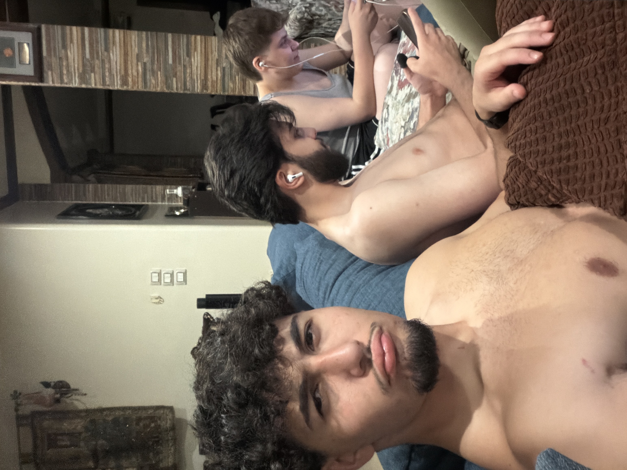 The media file is a vertically oriented photograph taken indoors, likely in a living room. Three young men are the primary subjects, all appearing to be in their late teens or early twenties. They are casually dressed and are situated on a couch, suggesting a relaxed social gathering.
The man in the foreground is closest to the camera, his face and upper chest filling a significant portion of the frame. He has dark, curly hair and a goatee, and his expression is neutral, with slightly pursed lips. He is shirtless. Behind him, another young man is also shirtless, seen in profile, with dark hair and a beard. He is wearing earbuds and appears to be looking at something off-camera. The third individual, positioned further back, is wearing a grey tank top and is also looking away from the camera, with short, light brown hair and earbuds in place. He is holding a smartphone.
The setting includes a couch with blue and patterned upholstery, and a textured brown throw blanket draped over the back. In the background, there are shelves with some decorative items, including a framed picture. A wall with light switches is also visible. The lighting suggests it is daytime, but the indoor setting makes it difficult to ascertain the specific time or weather. The overall mood is informal and relaxed, characteristic of friends spending time together at home. There is no visible text in the image.