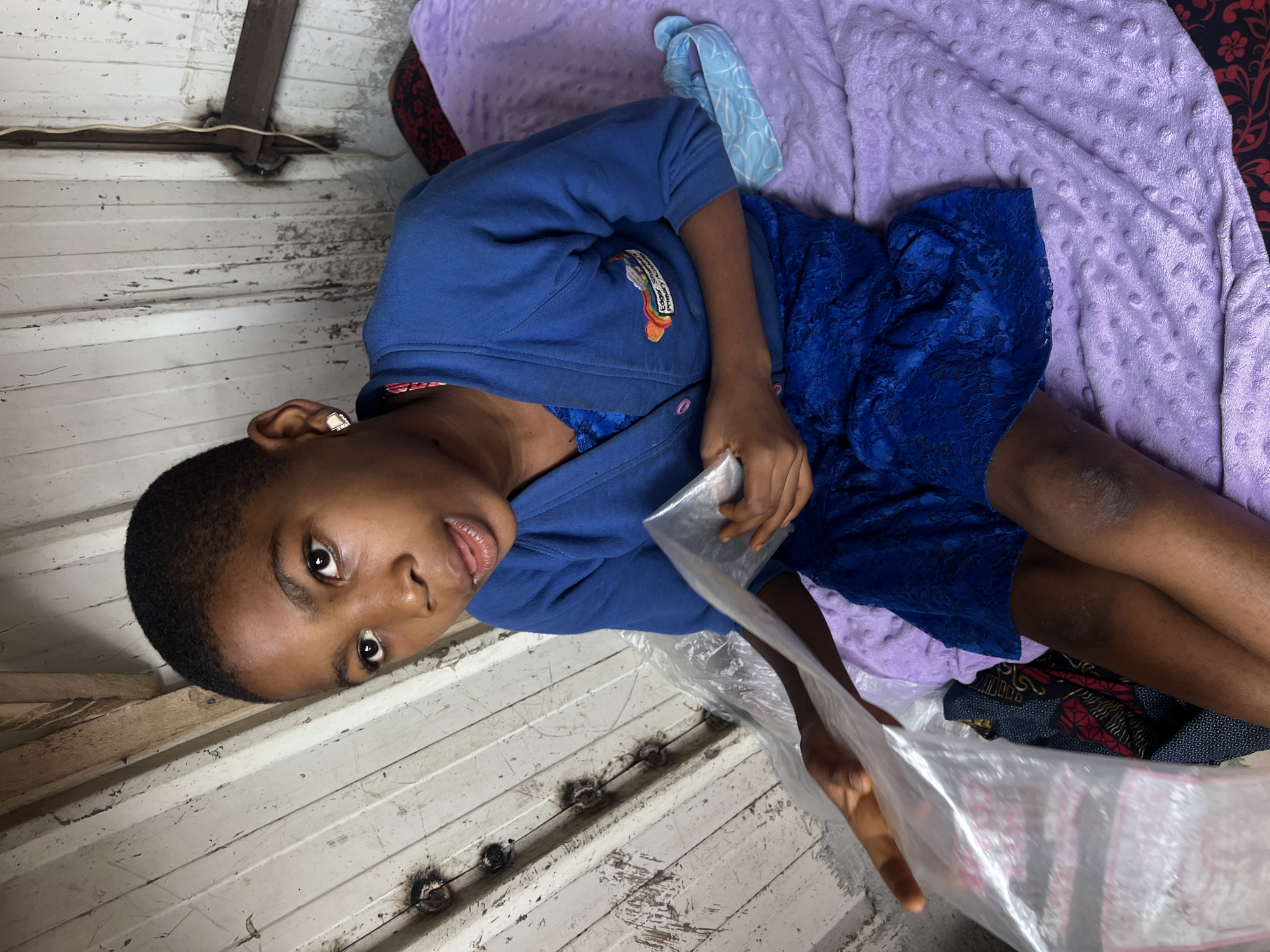 A young Black girl with short hair and an earring sits on a purple textured blanket in a room with corrugated metal walls. She is wearing a blue cardigan over a blue lace dress. Her left hand holds a clear plastic bag, and her right hand is visible near it. The girl looks towards the viewer with her mouth slightly open. The background shows the rough texture of the metal walls, with some visible nails and wires. The overall scene appears to be indoors, possibly in a makeshift or simple dwelling. The location is identified as Nkpor, Nigeria.