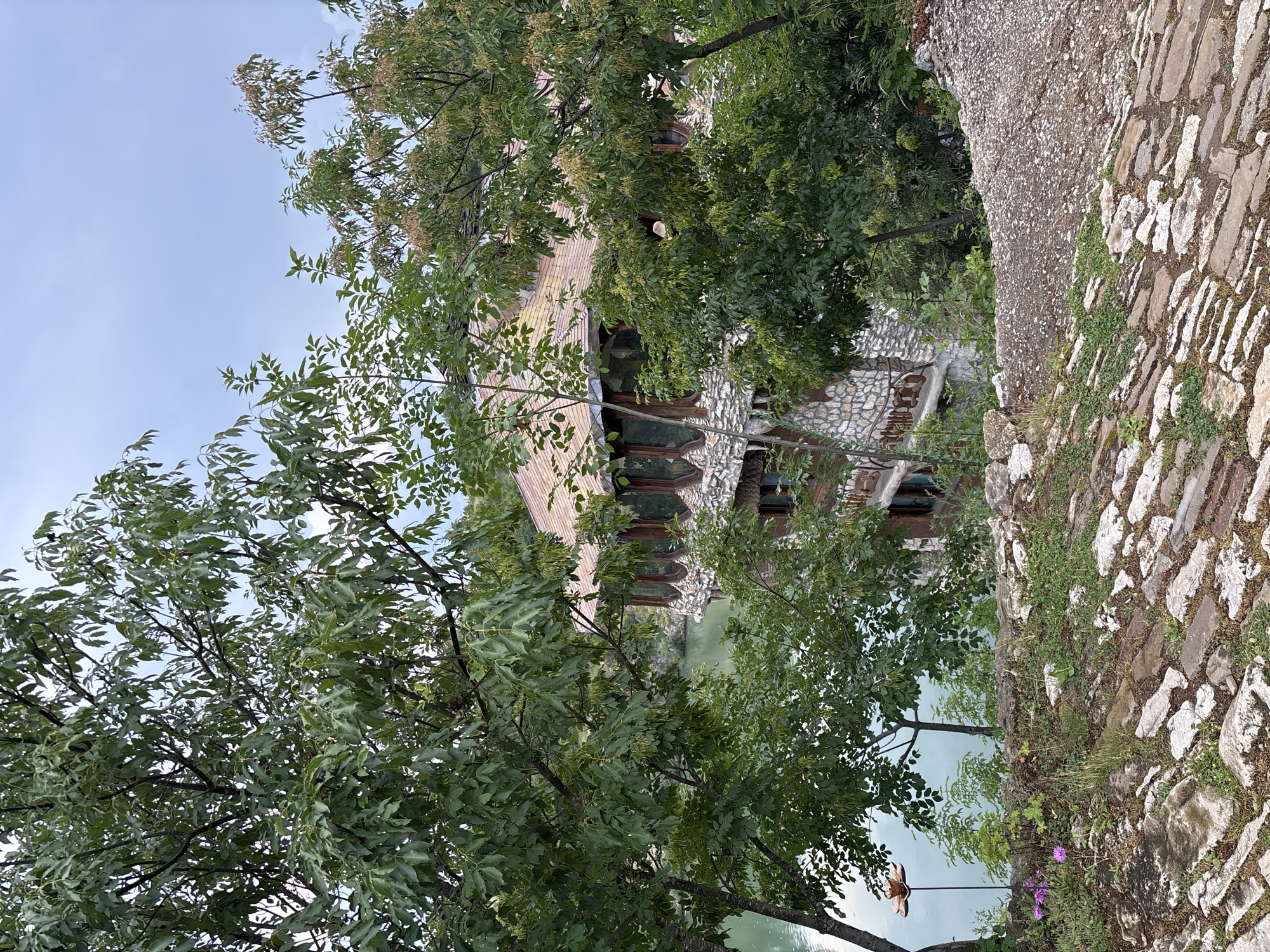 This detailed outdoor photograph, taken in Lukovit, Bulgaria, presents a multi-story building positioned beside a body of light green water, viewed through dense deciduous tree foliage from an elevated perspective. The foreground consists of an uneven path paved with rough, light-colored stones interspersed with green grass and small weeds. A cluster of small purple flowers is visible on the left, near a thin metal stake topped with a decorative, flower-shaped metallic object. This path transitions into a coarser, gravelly ground surface towards the right.
The building, partially obscured by trees, features a sturdy lower section constructed from irregular grey and brown stone blocks. Its upper visible level includes a series of distinctive, curved windows with dark wooden frames, offering expansive views of the adjacent water. The roof is complex, tiled with weathered reddish-brown and orange tiles, and appears to have multiple gables or dormer structures. The calm water, a pale green or turquoise hue, reflects the surrounding environment. Various trees with vibrant green leaves, some exhibiting distinctive seed pods, frame the scene, with one prominent tree on the left foreground. The sky above is uniformly overcast and light blue-grey. No human figures are present in the image.