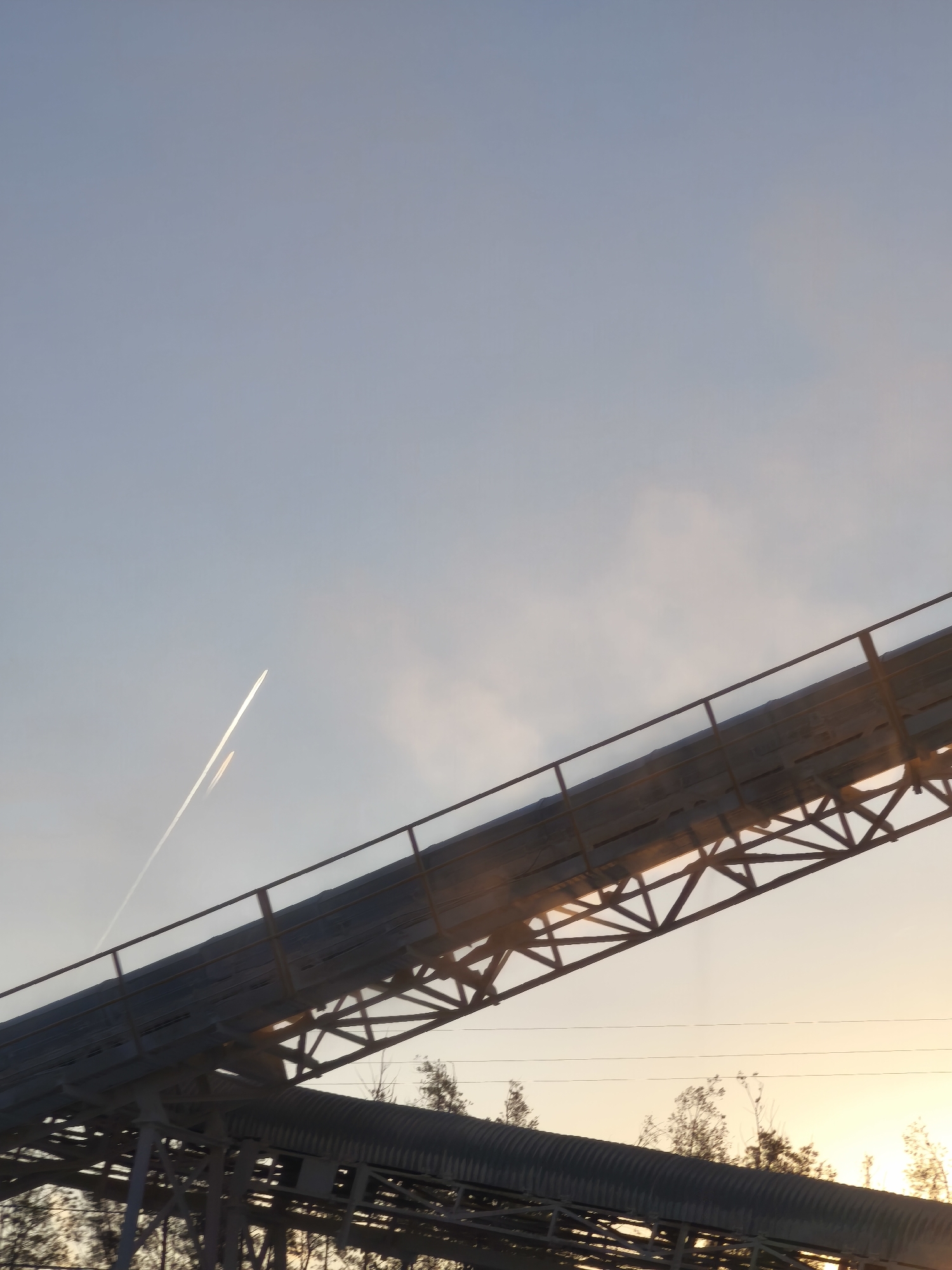 The image depicts an industrial conveyor belt structure in Riba-roja de Túria, Spain, seen against a backdrop of a light blue sky. The sky shows an airplane contrail. The image captures a clear, industrial scene under daylight.