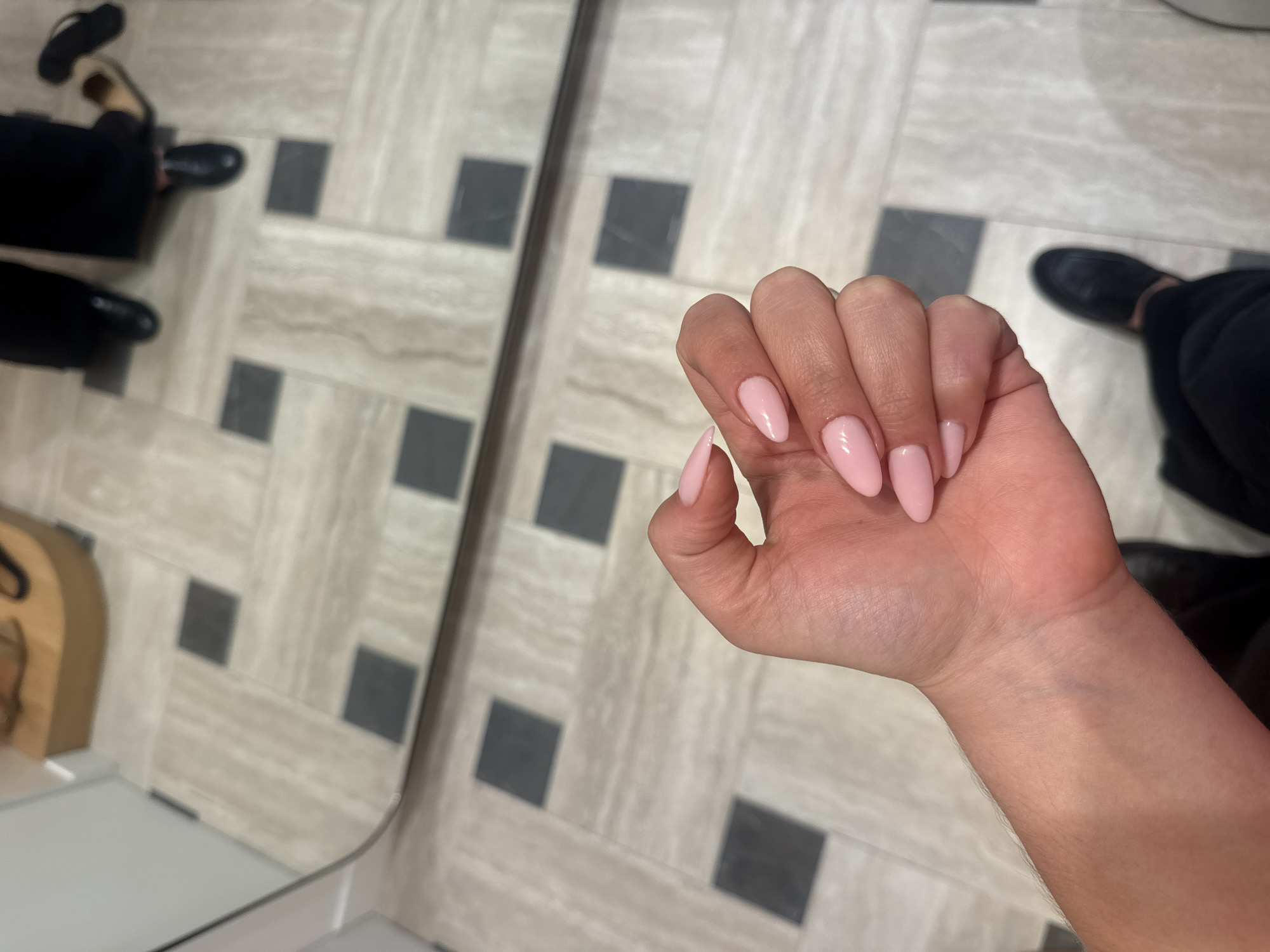 A person's hand with light pink, almond-shaped nails is prominently displayed in the center of the frame. The hand is held against a background of a mirror reflecting a tiled floor with a geometric pattern of light and dark squares. Reflected in the mirror are parts of two individuals dressed in dark clothing and black shoes, standing on the tiled floor. One individual appears to be wearing sandals and black clothing, while the other is wearing black shoes. The environment appears to be indoors, with neutral lighting. There is no information to support Paris, France as a location.