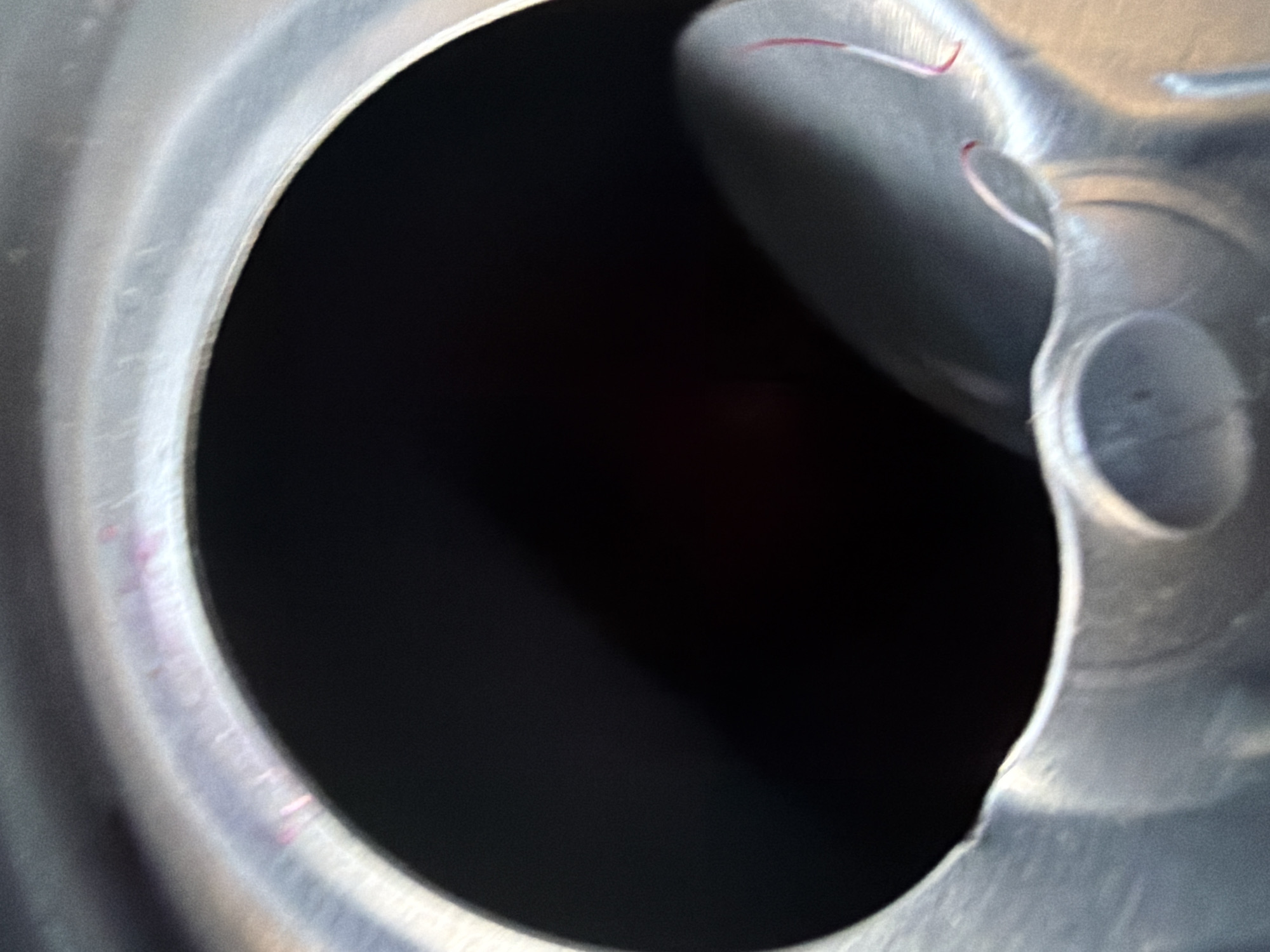 This is a close-up, overhead view of the interior of an open metal can. The dominant features are a large, dark, circular opening, likely the mouth of the can, which takes up the majority of the frame. The interior of the can appears to be dark, possibly reflecting a dark liquid or shadow. Surrounding the opening is the metallic rim of the can, which shows some subtle color variations, including faint pinkish or purplish streaks. To the right of the opening, a curved section of the can's interior wall is visible, with a small, flat piece of what appears to be plastic or metal detached and resting on the surface. This detached piece has a thin red thread or fiber adhered to it. The overall impression is of a utilitarian object, possibly a food or beverage container. There is no indication of the city of Kareby, Sweden, or any environmental context.