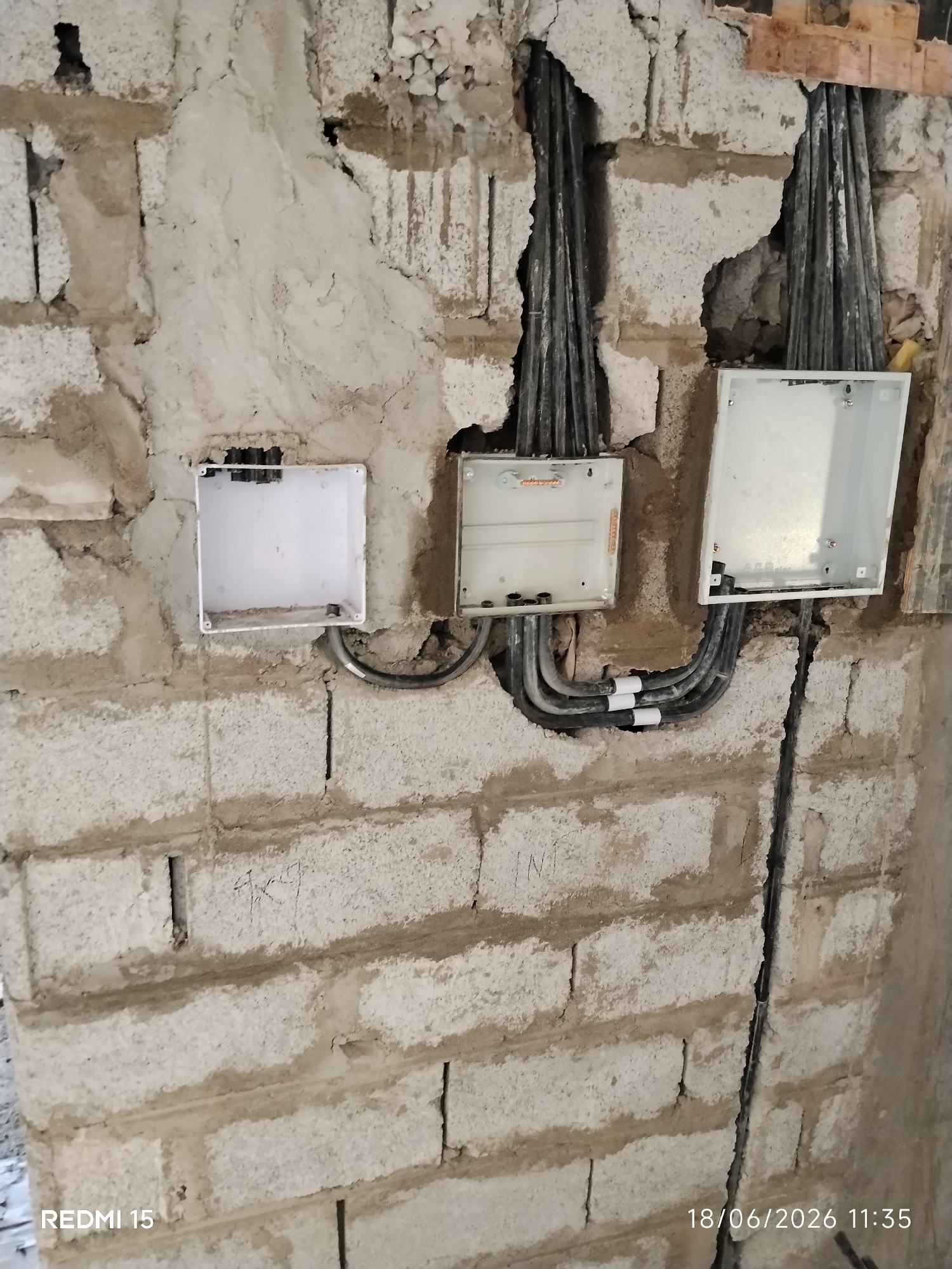 The image depicts an unfinished construction site wall in Unguwar Rimi, Nigeria, with electrical conduits and junction boxes installed. The wall is constructed from light-colored cinder blocks, some of which are partially covered with a rough plaster. Three white junction boxes are visible, two open and one closed. The two open boxes are situated on the right side of the image, with electrical cables entering from above and exiting downwards. The closed box, on the left, is also open, revealing an empty interior. Several black electrical conduits, bundled together, are emerging from the wall and entering the junction boxes. Some of the conduits have white tape wrapped around them. The overall environment appears dusty and unrefined, indicative of ongoing construction work. The bottom left corner of the image displays the text "REDMI 15", and the bottom right corner shows a date and time stamp: "18/06/2026 11:35". There are no people or visible signs of recent human activity, beyond the installation of the electrical components.