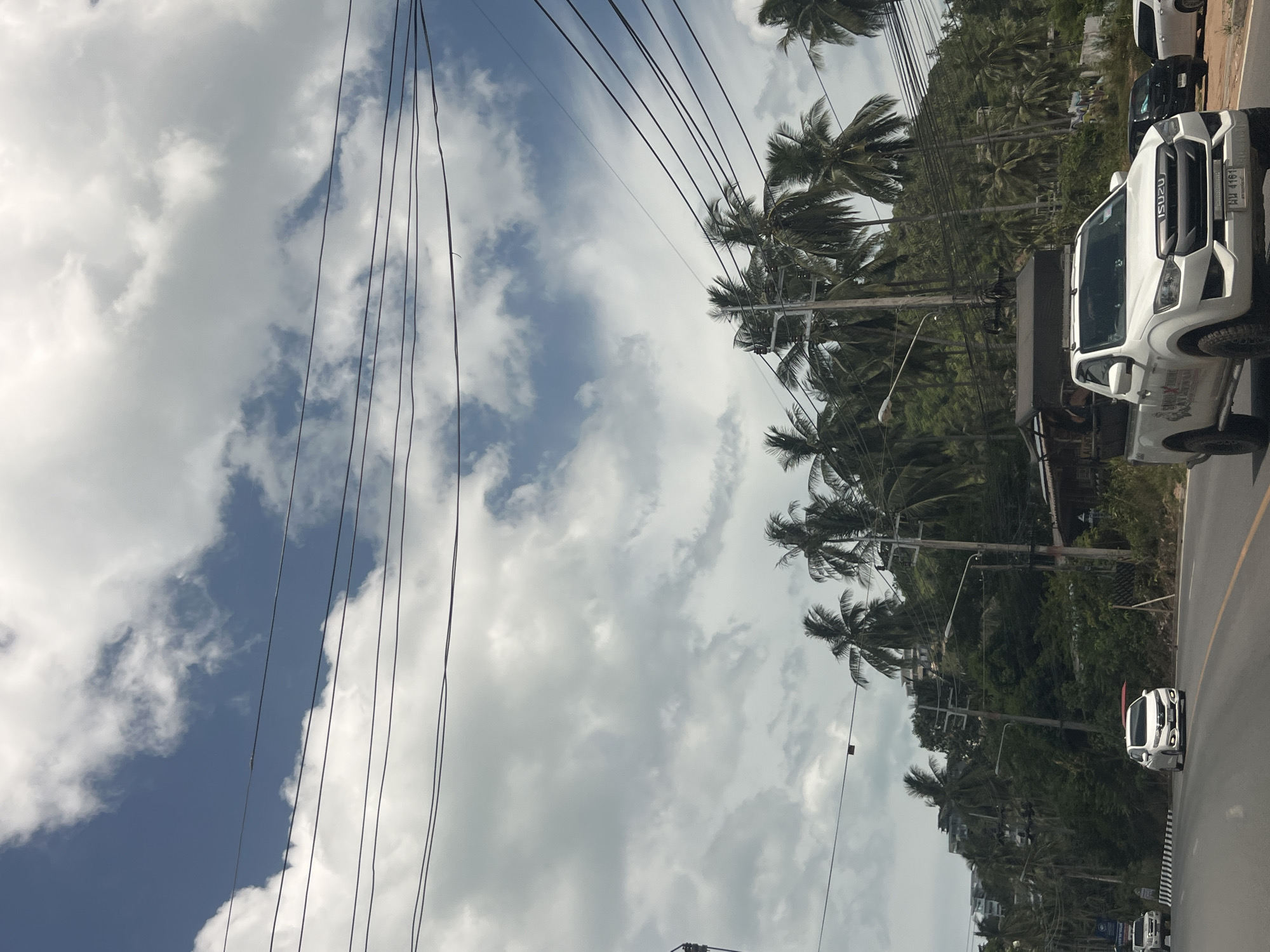 In the city of Ko Samui District, Thailand, a white pickup truck is captured in motion on a paved road. The truck, adorned with decals and sporting a license plate that reads "พล 4161", has a dark-colored canopy on its bed. Ahead of it, a white sedan is also traveling. The road is bordered by lush greenery and a dense growth of palm trees, indicative of a tropical environment. Overhead, a network of power lines crisscrosses the frame against a backdrop of a partially cloudy blue sky. The scene appears to be a typical daytime street view in a coastal or hilly area of the island.
