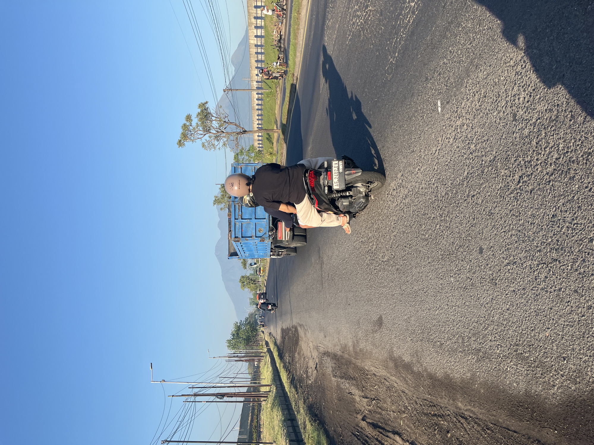 The image depicts a street scene in Siring, Indonesia, under a clear blue sky. In the foreground, two people are riding a motorcycle, with the passenger in the back wearing a beige helmet and a black t-shirt. The motorcycle is moving on an asphalt road, and its license plate is visible, reading "AE 2402 EX". A large blue dump truck is traveling ahead of the motorcycle. In the background, several other vehicles, including cars and motorcycles, are visible on the road. Mountains form a distant backdrop, under which utility poles and power lines stretch along the side of the road. The environment appears to be rural or semi-urban, with vegetation and a ditch alongside the road. The time of day is likely morning or late afternoon, given the long shadows cast by the vehicles.
