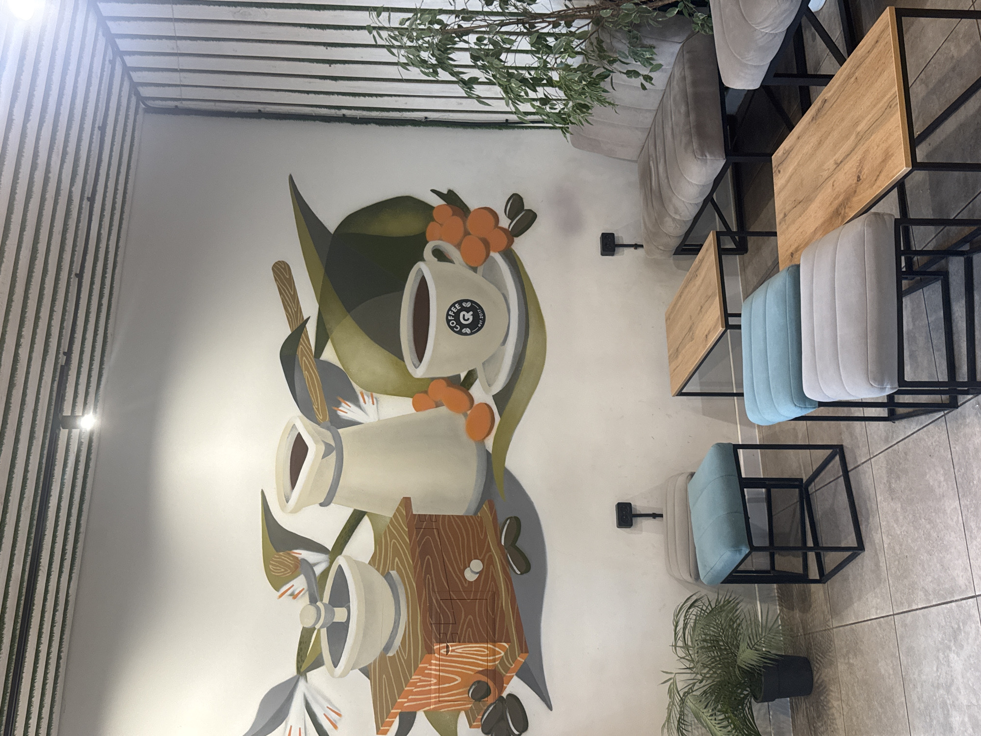 An indoor scene depicts a coffee shop with a large mural on the wall. The mural features a stylized illustration of coffee-making elements, including a coffee grinder, a coffee pot, a cup of coffee, coffee beans, and coffee cherries, all set against a backdrop of abstract green leaves and white space. In the foreground, there are several modern seating arrangements. These include plush, tufted seating in muted gray tones, and cube-shaped stools with teal and gray cushions, all supported by black metal frames. A small potted plant with green leaves sits in a black pot on the tiled floor. A narrow, wooden coffee table with a black metal frame is positioned in front of one of the seating areas. The lighting fixtures are visible on the ceiling, and the walls are painted white. The location is identified as a coffee shop in Chaikovsky, Russia.