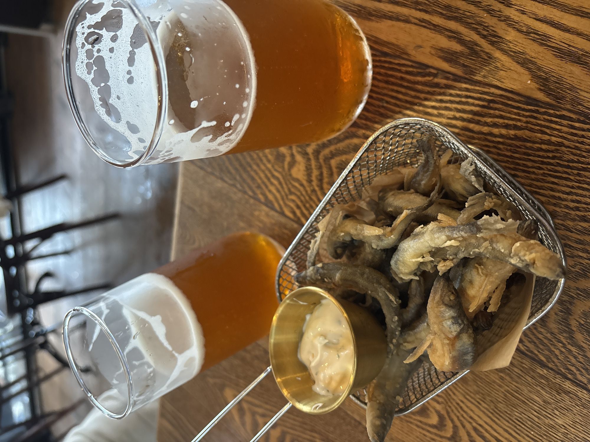 Two glasses of amber-colored beer, one nearly empty and the other partially full with a foamy head, sit on a dark wooden table. Next to the glasses is a small wire basket filled with fried fish, their crispy exteriors golden brown. A tiny brass cup holding a white dipping sauce sits in front of the basket. The scene is set indoors, likely in a restaurant or pub, given the table setting and the presence of drinks and food. The lighting suggests daytime, possibly late afternoon or early evening, as the light is warm and diffused. The overall mood is casual and relaxed, perhaps a meal being enjoyed with a drink. No text is visible in the image.