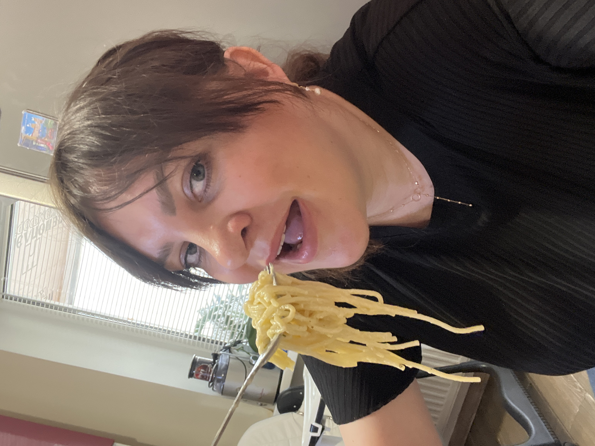 A woman with short brown hair is holding a fork of spaghetti up to her mouth. She is wearing a black shirt and a delicate necklace. The background includes a window with blinds, a small plant, and a kitchen appliance. The image is taken at an angle. There is no indication the location is Noviy, Russia.