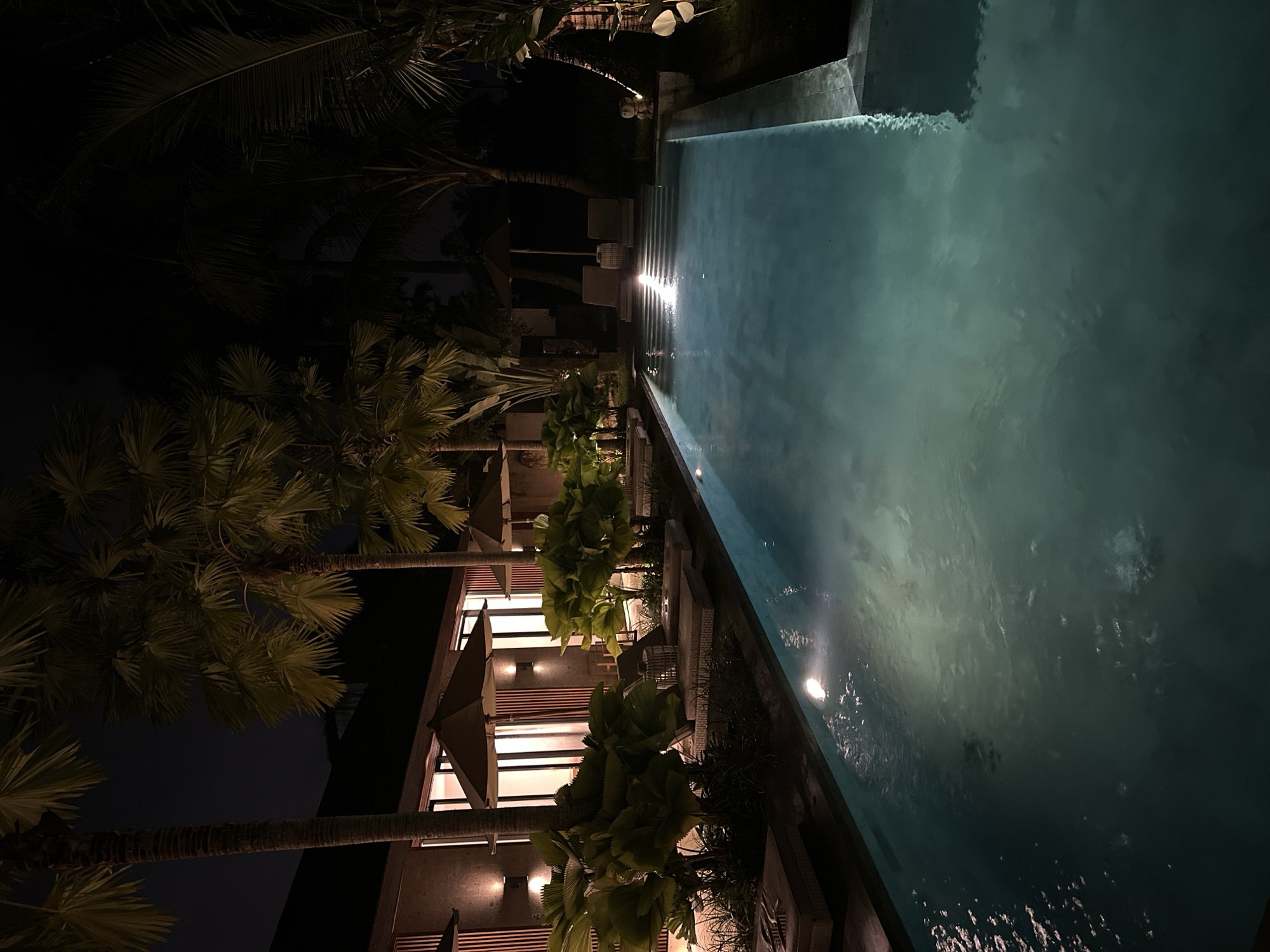 Here is a detailed description of the image:
The image depicts a dimly lit outdoor scene at night, possibly in Sukawati, Indonesia, given the mention of the location. The main focus is a swimming pool stretching out into the dark background. The water's surface reflects the dim light.
On the left side of the frame, a building is visible, illuminated by warm interior lights shining through windows covered with horizontal slats. Umbrellas and palm trees line the area between the building and the pool, adding to the tropical ambiance. The sky is dark, indicating that it's nighttime.