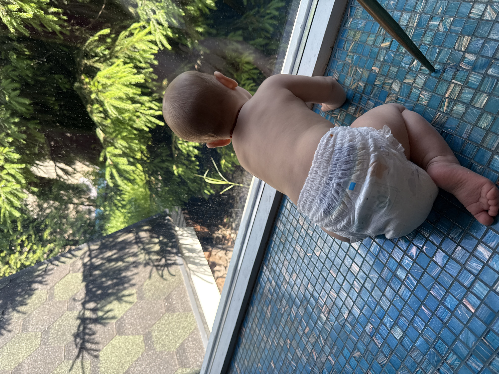 A baby wearing a diaper lies on their stomach on a blue tiled surface near a window. The baby is looking out the window at a green, leafy tree. Outside the window, the ground is covered in patterned tiles with shadows from the tree. A table leg is also visible on the tiled surface. The location of the image is Чёрное, Russia.