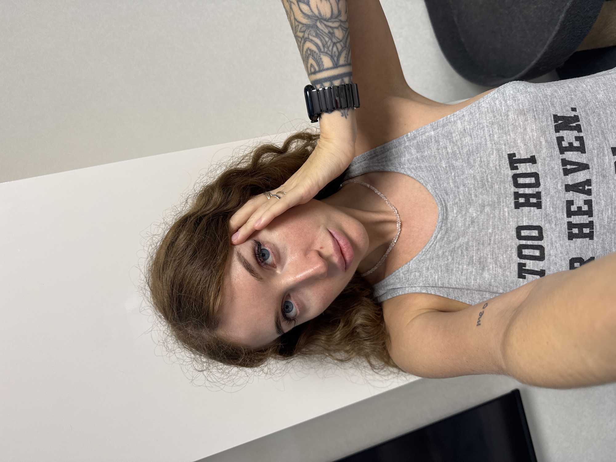 The image is a selfie depicting an adult female indoors. She has long, wavy, reddish-brown hair and blue eyes, with a small stud piercing on her right nostril. Her left hand is resting against her forehead, with a silver-toned ring on her ring finger. She is wearing a grey tank top with black text partially visible on the chest, which reads "TOO HOT FOR HEAVEN." A delicate, light-colored beaded necklace is around her neck. On her left inner bicep, a small black tattoo spells "me o." Her right forearm features a large, intricate black and grey tattoo of a mandala or floral design. On her right wrist, she wears a black Apple Watch with a segmented black link bracelet. The background consists of plain, light-colored walls, with a vertical white panel visible behind her left shoulder and a dark rectangular object partially visible on the far left. A dark, circular, textured object is partially visible on the far right. The image does not contain any visually discernible landmarks, signage, or architectural details that would allow for geo-localization to the city of Chaikovsky, Russia, or any specific geographic location.