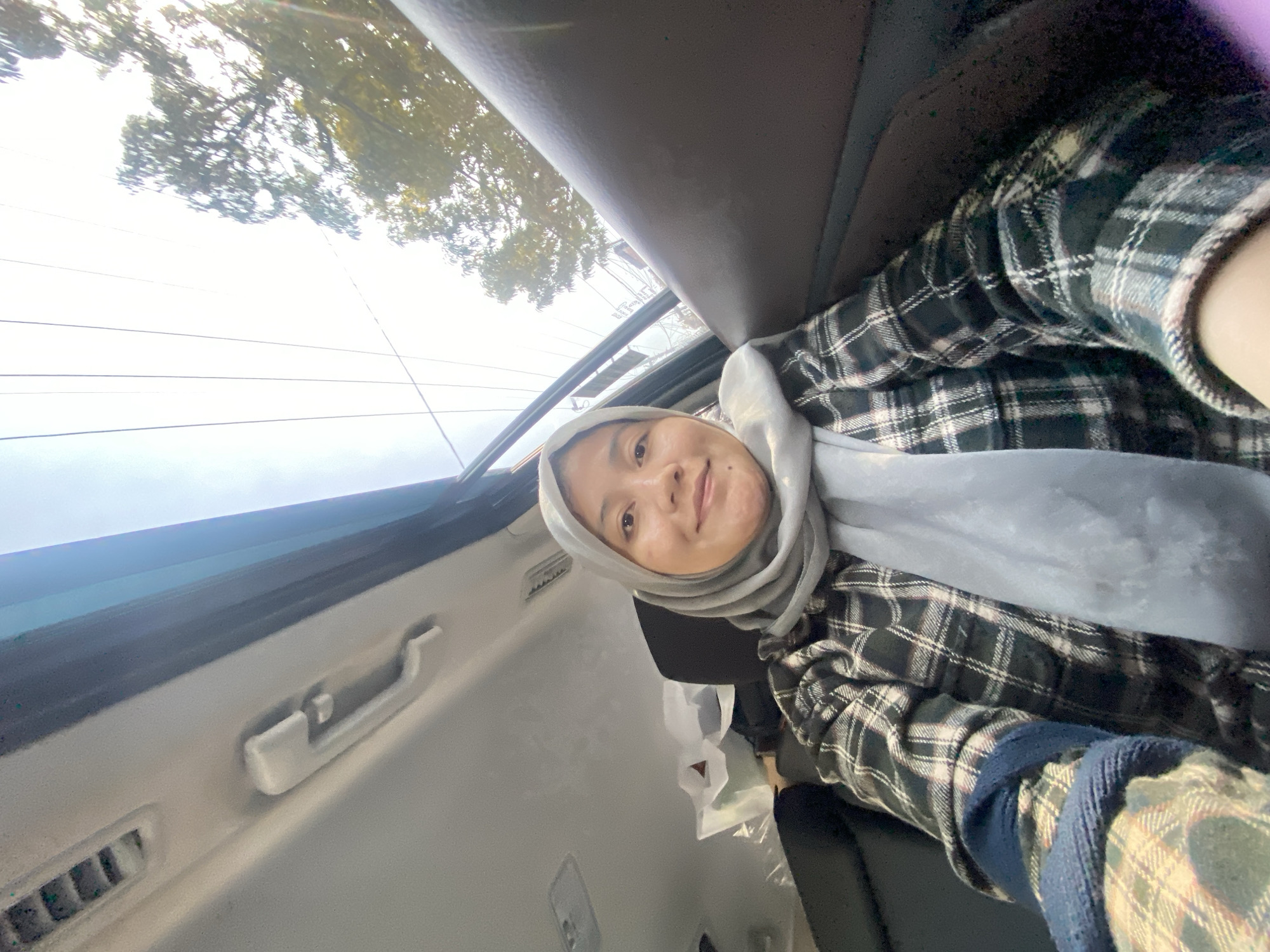 A woman wearing a gray hijab and a plaid shirt is sitting in the passenger seat of a car, looking at the camera. The car's interior is visible, including the headliner and door panel. Outside the car, trees and power lines are visible against a bright sky. The scene appears to be in a residential area of Banjarmasin, Indonesia.