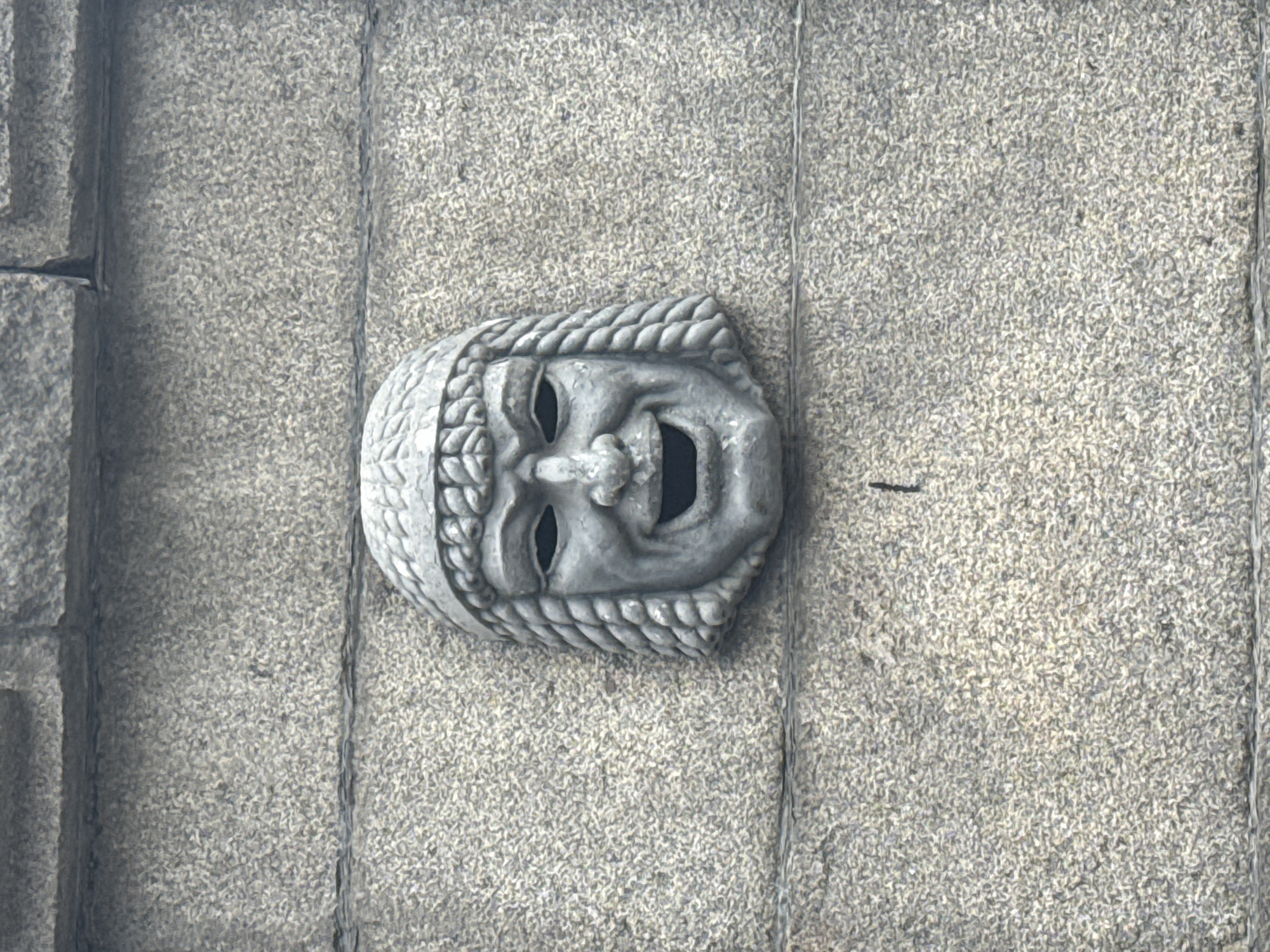 An ornate grey stone mask, depicting a broadly smiling or laughing face with an open mouth and prominent eyeholes, is set into a textured grey stone wall. The mask features a detailed, braided or patterned border resembling hair or a headpiece. The surrounding wall is constructed from large blocks of coarse-grained grey stone, possibly granite, with clearly visible vertical and horizontal joints.
The mask itself is a static architectural embellishment, likely part of a building facade, and evokes the style of a classical comedic theatrical mask. No specific activity or event is taking place; it is a fixed decorative element. The lighting suggests ambient daylight, creating soft shadows that accentuate the relief carving of the mask and the rough texture of the stone. There are no notable cues regarding the time of day or specific weather conditions other than the presence of daylight. No visible text is present in the image. This architectural detail is found in Saint Petersburg, Russia.