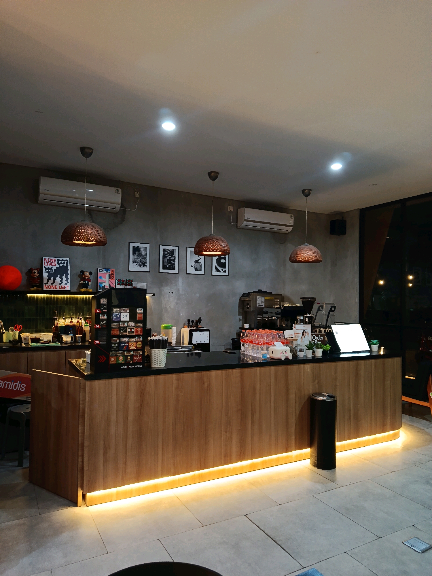 The image depicts the interior of a coffee shop in Mangunjiwan, Indonesia. The counter is made of wood and is illuminated by warm LED lights. Behind the counter, there are various items, including a coffee machine, bottles of drinks, and decorative items. The walls are gray concrete and decorated with framed pictures. There are also hanging lamps and an air conditioning unit visible. The floor is tiled. The scene is well-lit, creating a cozy atmosphere.
