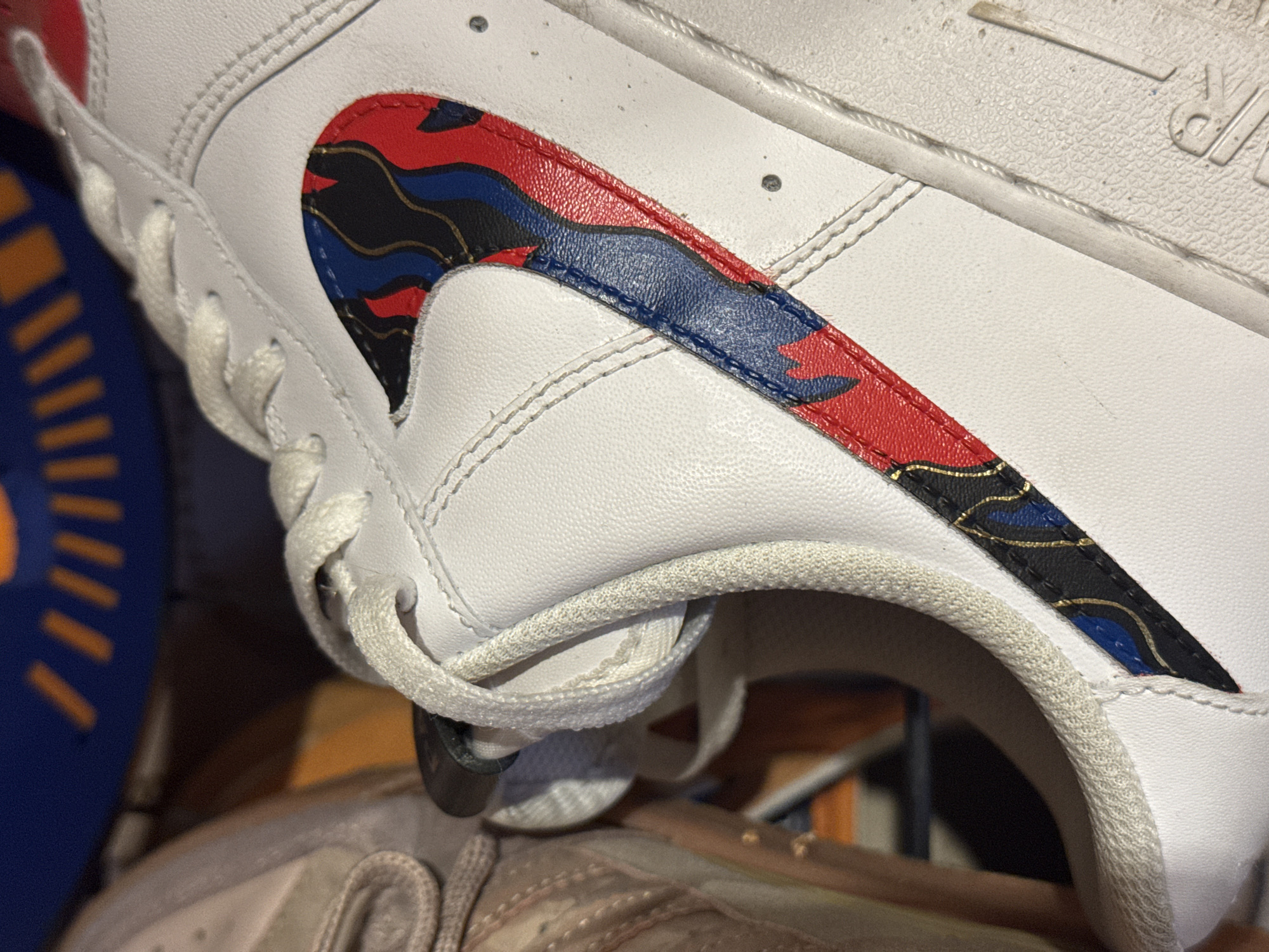 A close-up image shows the side of a white leather sneaker. The iconic "swoosh" logo is present on the side, but it is filled with a complex, patterned design in red, blue, black and gold. The laces of the shoe are white and knotted. Portions of other objects (likely other shoes) are visible in the background, with one appearing blue and another a lighter, brownish-grey. The lighting suggests indoor conditions. The location is not determinable from the image.