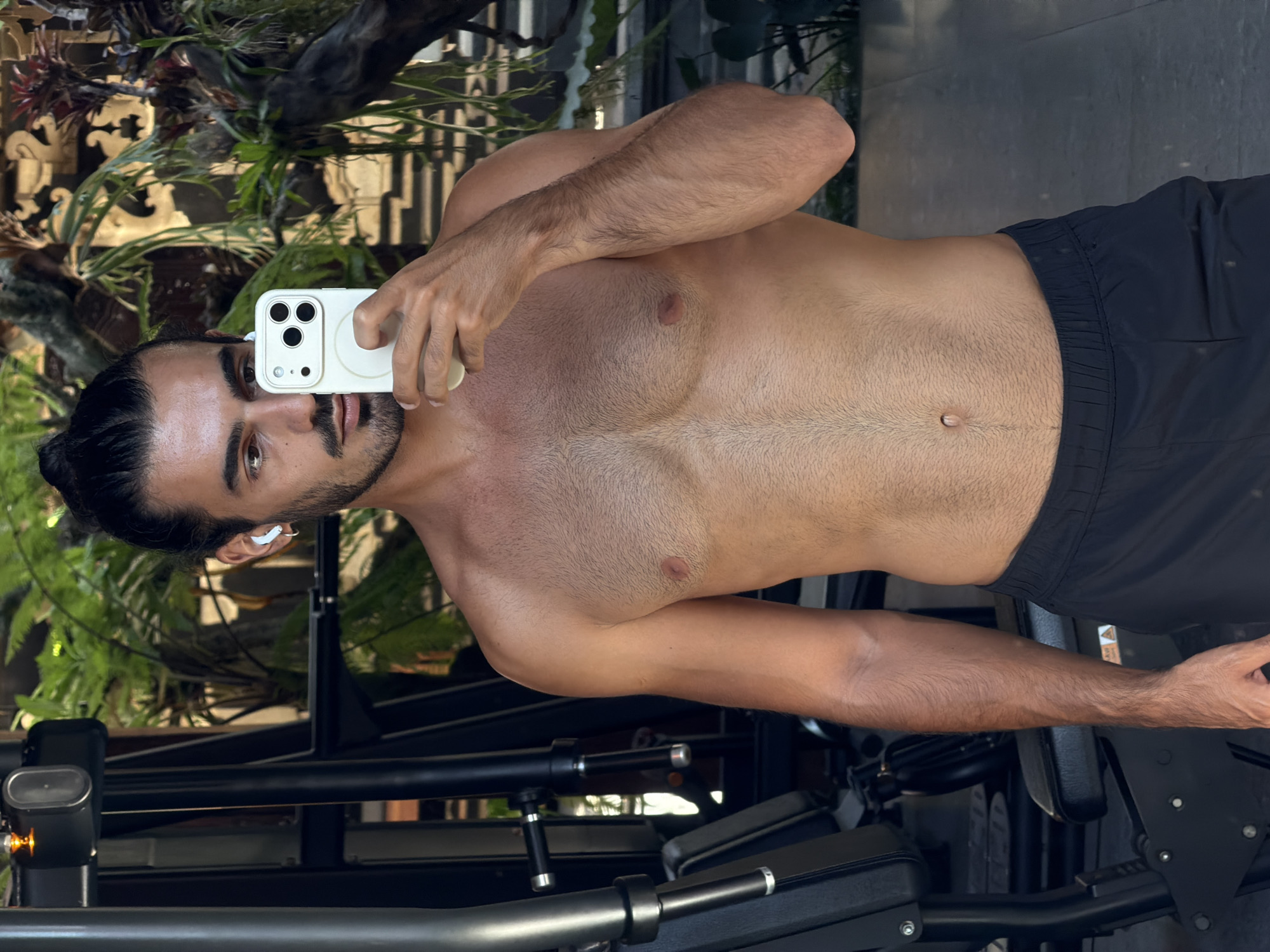 A shirtless man with dark hair pulled back in a bun, a trimmed beard, and an earbud in his left ear is holding a white smartphone in his right hand, presumably taking a selfie. He is wearing black shorts. The background includes foliage and a structure that resembles a piece of decorative woodwork. Below him is a black piece of equipment that looks like part of a workout machine. It is daytime, and the photo was taken in North Kuta, Indonesia.
