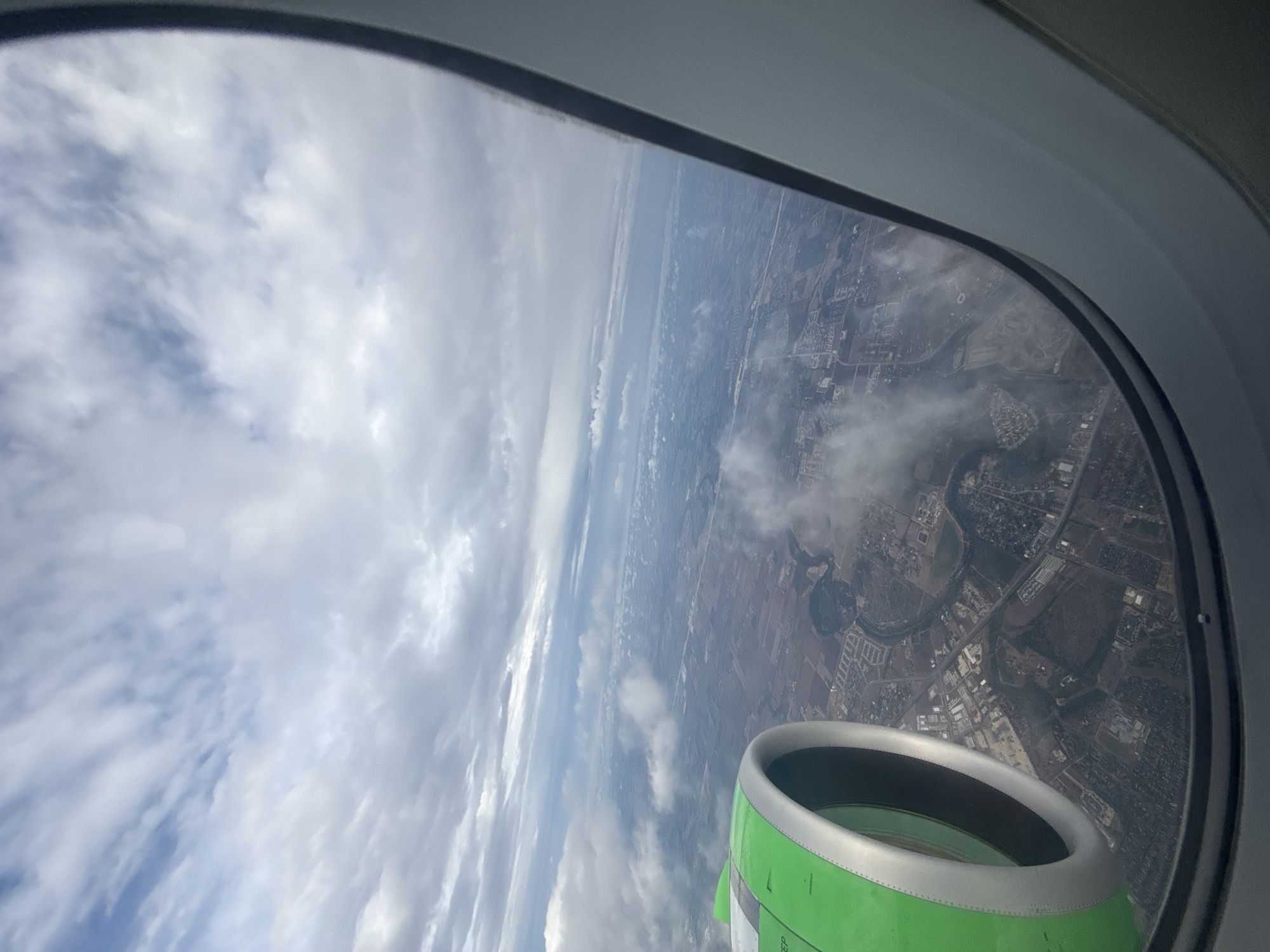 The view from an airplane window shows a cloudy sky on the left and a landscape of Schertz, Texas, on the right. The landscape includes a river or creek winding through urban and rural areas, with buildings, roads, and fields visible. A green and silver jet engine is partially visible in the lower right corner of the window.