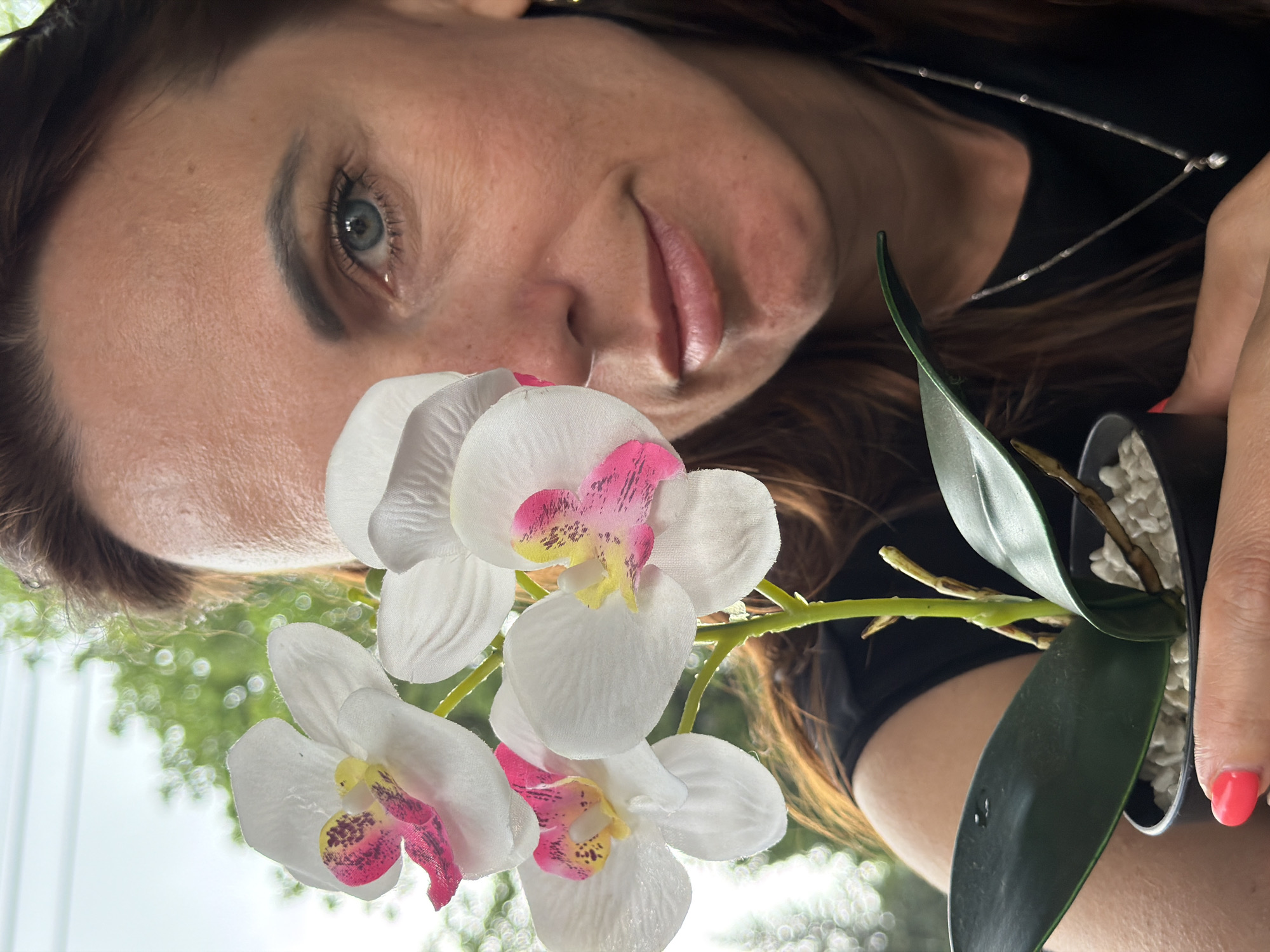 In this close-up, vertical, eye-level shot, a woman with brown hair and blue eyes holds a potted orchid plant in front of her face, obscuring most of her left side. She is wearing a black top and a delicate silver necklace. Her right eye is visible, looking directly at the camera. The orchid has white petals with pink and yellow accents. The background consists of blurred green foliage and a bright sky, suggesting an outdoor setting. The overall impression is a portrait with a floral element, possibly taken in a garden or on a balcony. The image does not contain any specific landmarks or elements that would indicate the city of Birobidzhan, Russia.