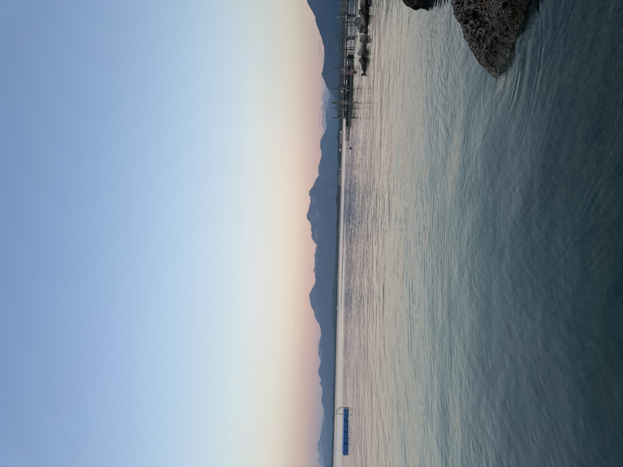 An expansive, calm body of water, likely Lake Chiemsee, dominates the foreground and middle ground. The water reflects the soft hues of a twilight sky, transitioning from a pale blue at the top to gentle pinks and oranges near the horizon. In the distance, a range of dark, silhouetted mountains forms a dramatic backdrop.
To the left of the frame, a blue floating platform with a white ladder is anchored in the water. Further to the right, a wooden pier extends out from the shore, with several structures and what appear to be boats moored alongside. The shoreline is marked by a few dark rocks in the lower right corner of the image, with one prominent rock emerging from the water.
The scene suggests a peaceful evening in Prien am Chiemsee, Germany, a town known for its picturesque location on the lake and its proximity to the Bavarian Alps. The clear sky and still water indicate favorable weather conditions. There are no people visible in the image.