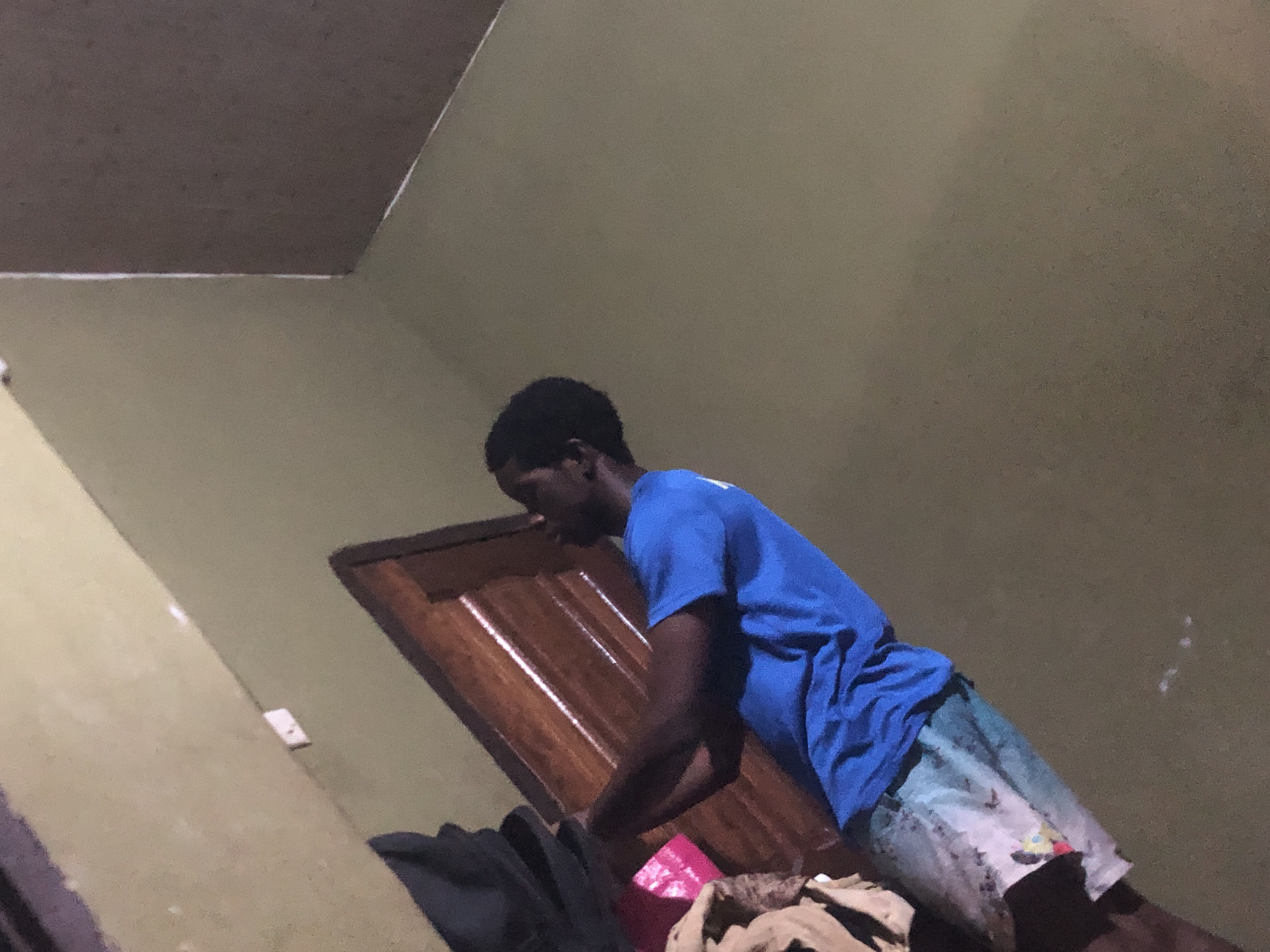 A dimly lit indoor scene features a dark-skinned man wearing a bright blue t-shirt and patterned shorts. He appears to be leaning over a pile of items. A brown wooden door with vertical panels is visible behind him. The room has olive-green walls and a dark brown ceiling. A light switch is visible on the wall. The location is Awkuzu, Nigeria.