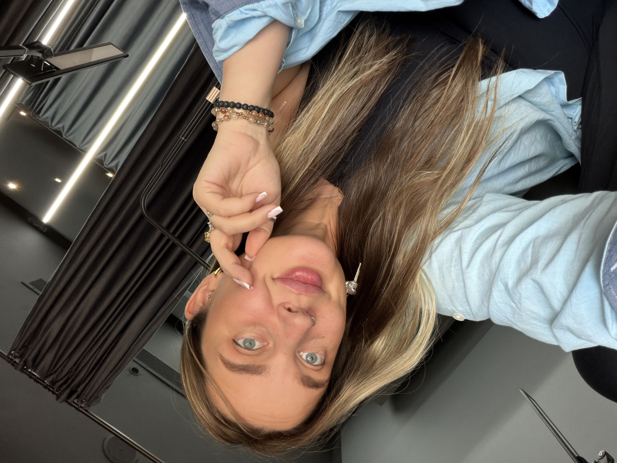 A young woman is facing the camera in what appears to be a selfie. She has light skin, blue eyes, and long, blonde hair with darker roots. She's wearing a light blue button-down shirt, black leggings, and bracelets on her left wrist. Her left hand is near her face. She has a nose ring and earrings. The background features black curtains, bright LED lighting, and gray walls and ceiling. There is no indication of this image being taken in Moscow, Russia.