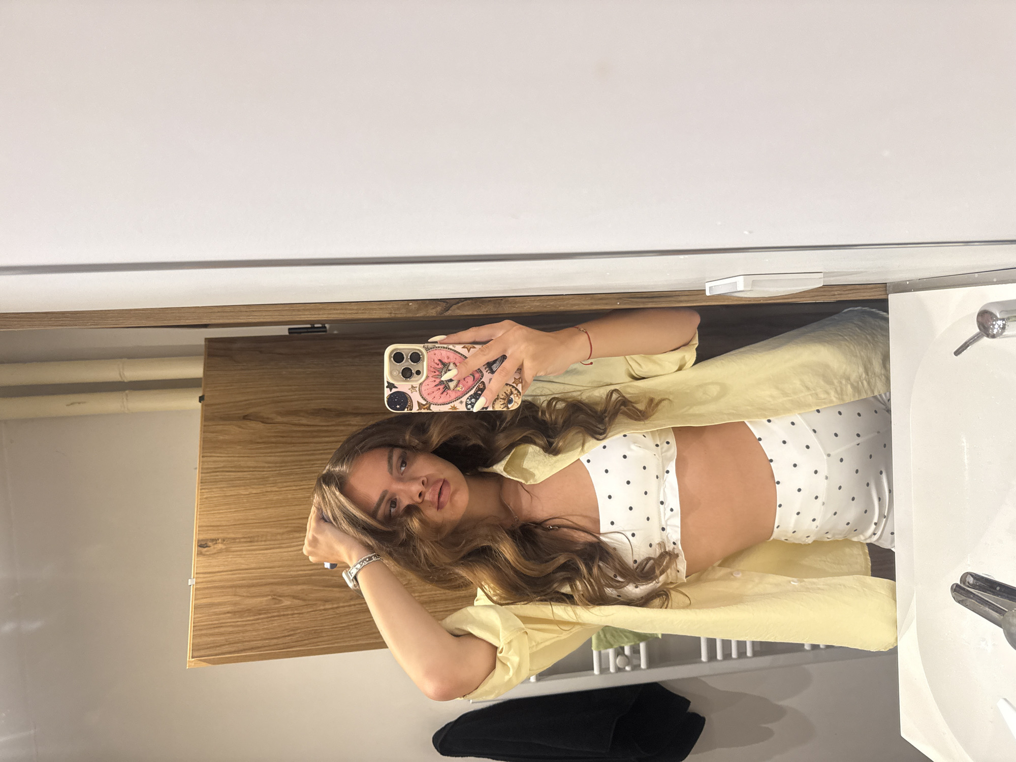 A young woman with long, wavy brown hair is taking a mirror selfie in a bathroom. She is wearing a white polka-dot bralette and matching polka-dot shorts, with an unbuttoned yellow shirt over the bralette. She has her left hand raised to her head and is holding her phone in her right hand. The bathroom features a wooden cabinet, a towel rack with white towels, and a white sink. The environment appears to be a modern residential bathroom, but there is no specific information to indicate it is located in Plovdiv, Bulgaria.