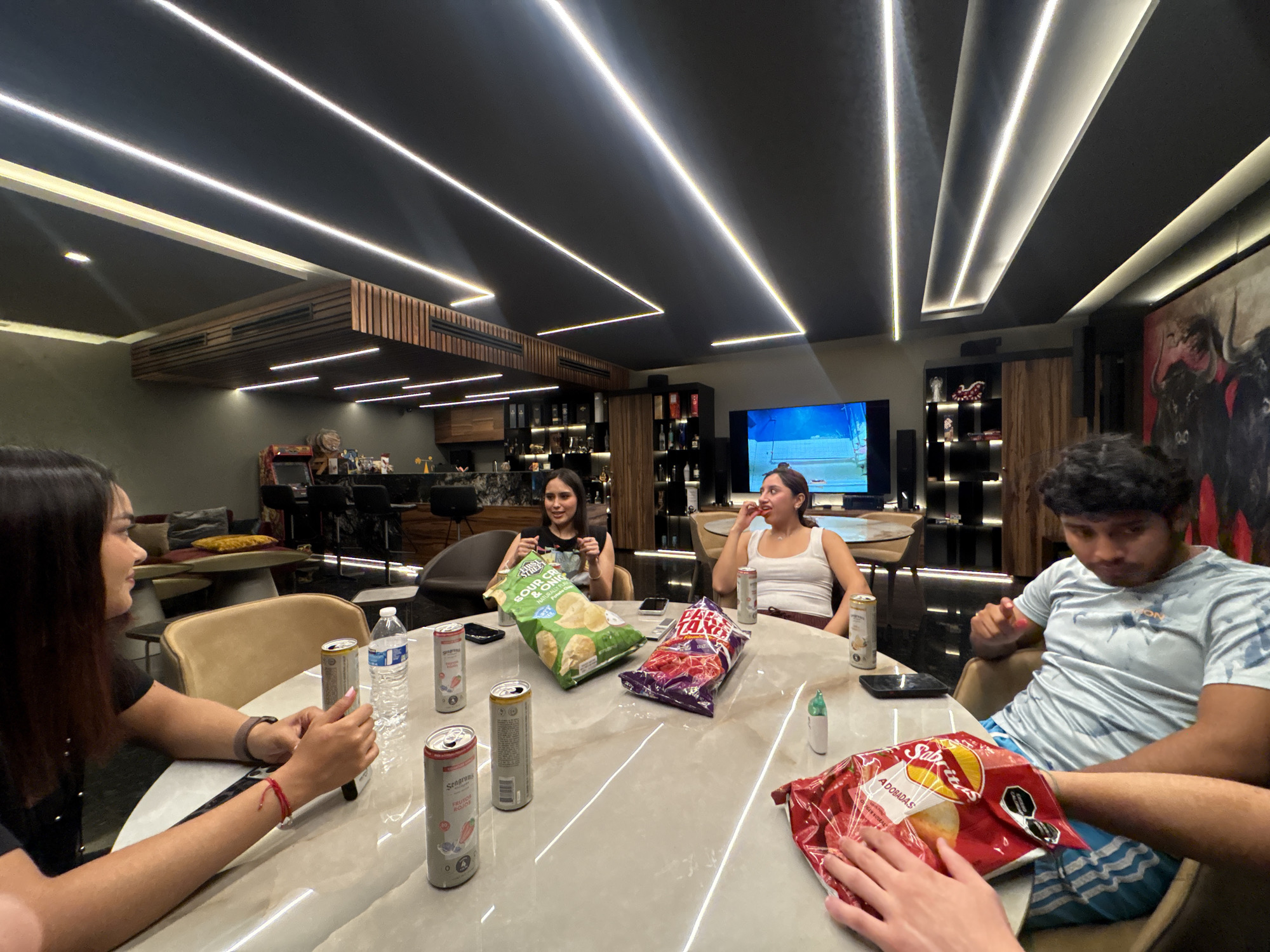 Four people sit around a round, marble-topped table. On the table are three open bags of potato chips (Sour Cream & Onion, Fritos Taxi, and Sabritas Adobadas), five cans of Seagram's Fruity Rose, and a bottle of water. A black mobile phone lies near the center of the table. The room features a black ceiling with parallel LED strip lights, a bar area in the background, a television, and art depicting bulls. The location is San Bernardino Tlaxcalancingo, Mexico.