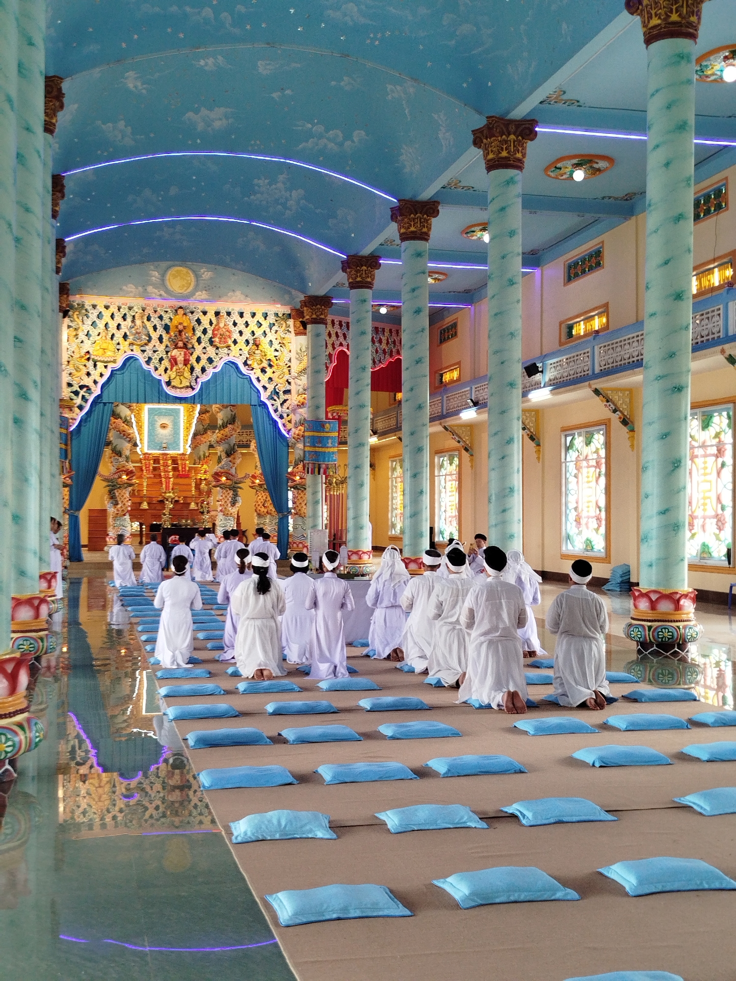 The image captures a religious ceremony inside a temple in Xuân Thành, Vietnam. Several individuals dressed in white robes are kneeling on a tan-colored floor covered with rows of small, light blue cushions. They appear to be in a state of prayer or meditation, facing an ornate altar at the far end of the hall. The altar is decorated with an intricate, blue-trimmed archway and statues. The temple's architecture features tall, light blue pillars with decorative tops, supporting a blue ceiling adorned with cloud-like designs and illuminated by soft, glowing lines. Stained-glass windows line the walls on the right. The floor is highly reflective, mirroring the scene.