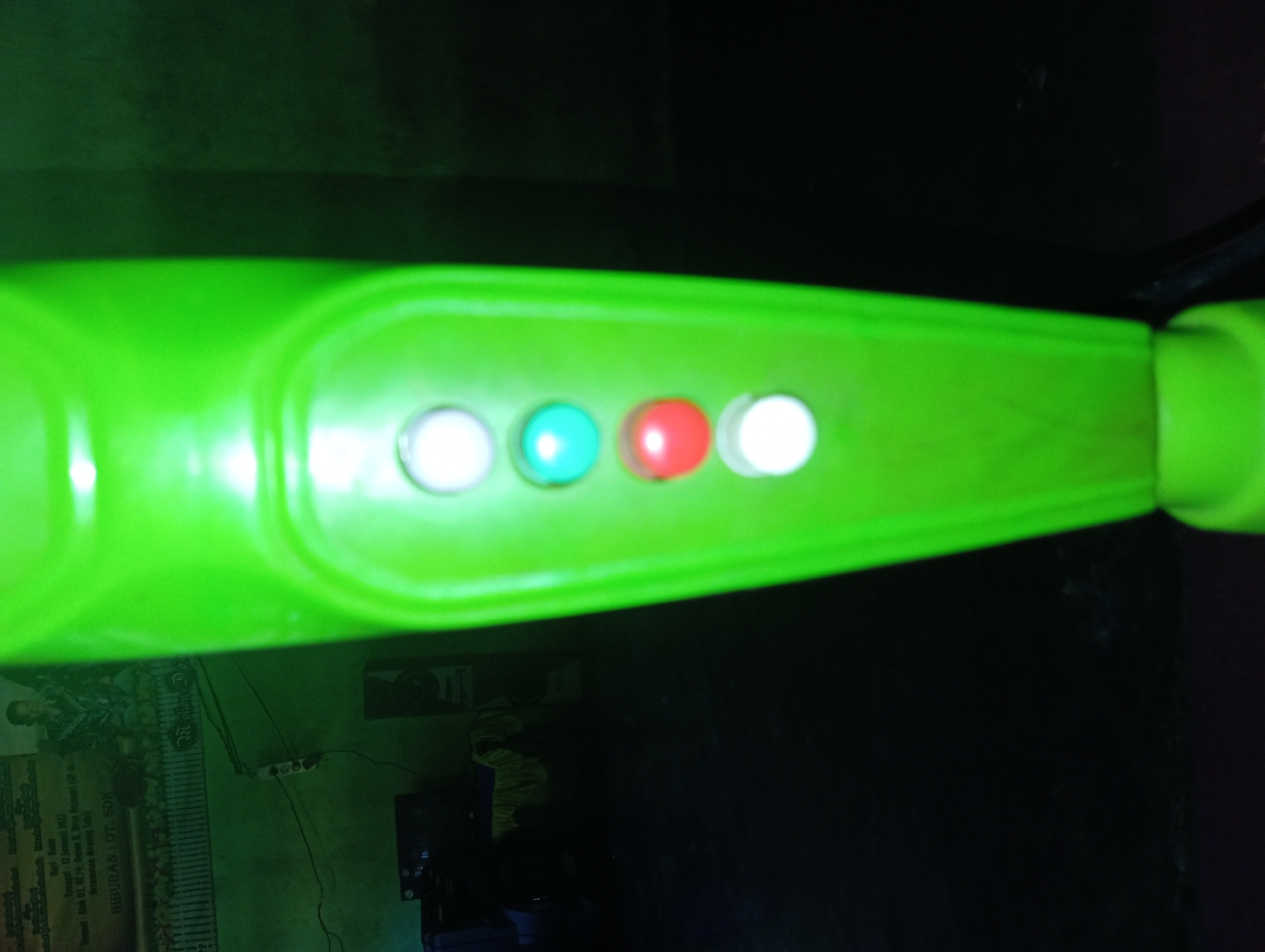 A close-up, low-angle shot focuses on the control panel of a bright green fan, positioned vertically in the foreground. The panel features a row of four circular buttons, two white, one turquoise, and one red, arranged from top to bottom. The background is dark and out of focus, but appears to show an indoor setting with a poster on the wall, potentially a calendar or informational notice, with Indonesian text. To the left of the fan, a dark electronic device, possibly a monitor or television, is visible, along with what appears to be a power strip plugged into the wall. The overall lighting is dim, creating a green hue on the fan and casting the background in shadow. The location is identified as Rejosari, Indonesia, suggesting an interior domestic or public space.