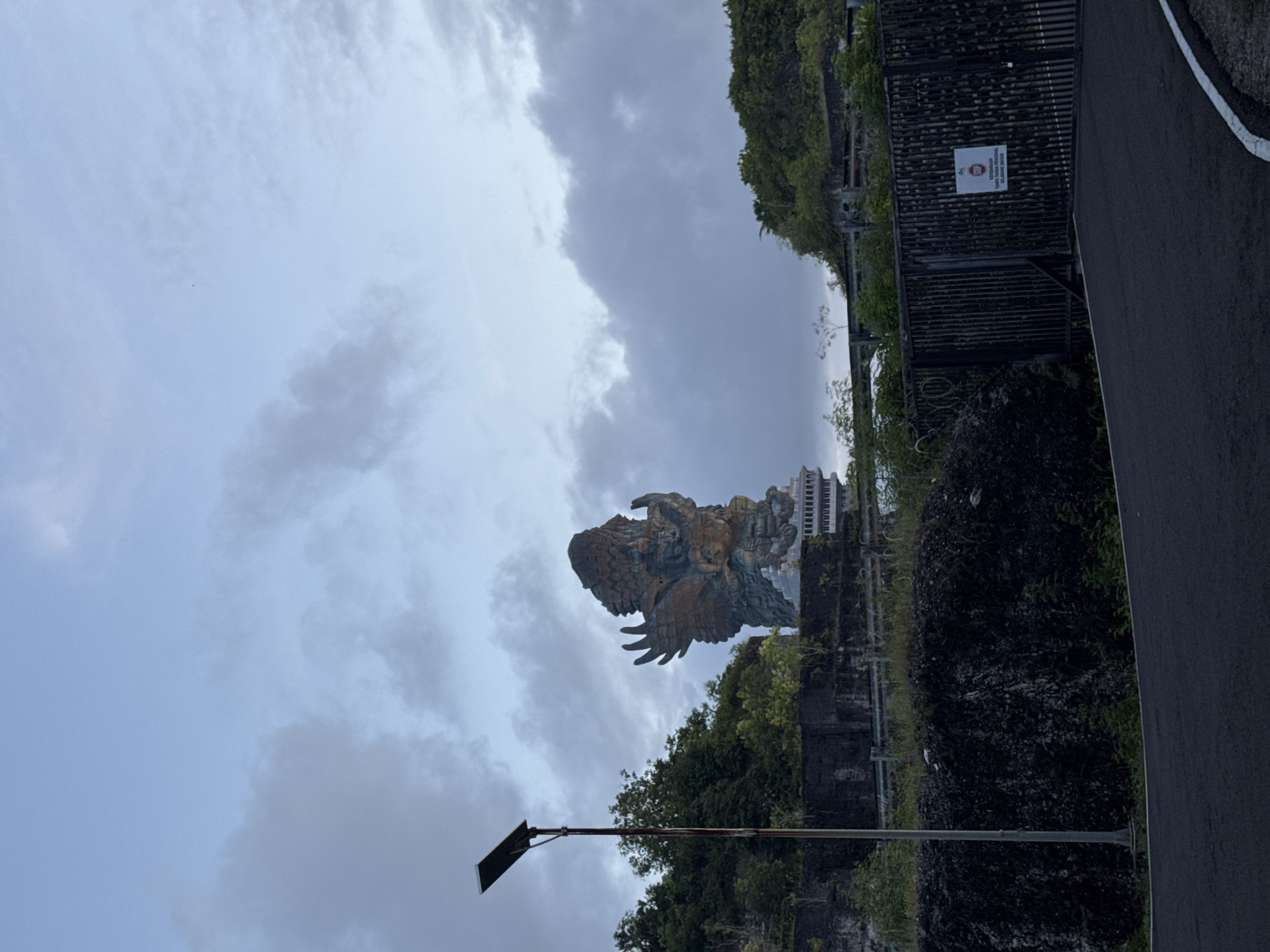The image presents a majestic outdoor scene featuring a monumental statue under an overcast sky. The central subject is a colossal, weathered statue, likely depicting the Hindu deity Vishnu mounted on the mythical eagle Garuda. The statue is intricately detailed, with broad wings and a powerful, regal form, rendered in bronze and greenish hues, suggesting age and grandeur.
The statue is situated on an elevated, rocky base covered with lush green vegetation, including dense trees and bushes. To the right of the statue, a paved asphalt road or pathway curves, bordered by a tall, dark metal fence or gate. Beyond the fence, more greenery is visible, and in the distance, a modern building with curved, multi-story architecture can be seen. A street light on a pole is visible in the lower-left foreground.
The sky is dominated by thick, grey clouds, indicating an overcast day with soft, diffused lighting. There is no specific human activity taking place.
A white rectangular sign with red lettering and a red "no entry" symbol is attached to the metal fence. The Indonesian text on the sign reads: "dilarang KENDARAAN ANGKUTAN BARANG DILARANG MASUK," which translates to "forbidden GOODS TRANSPORT VEHICLES PROHIBITED ENTRY." Based on the iconic statue and the location context of Kuta Selatan, Indonesia, the scene is identifiable as the Garuda Wisnu Kencana Cultural Park.