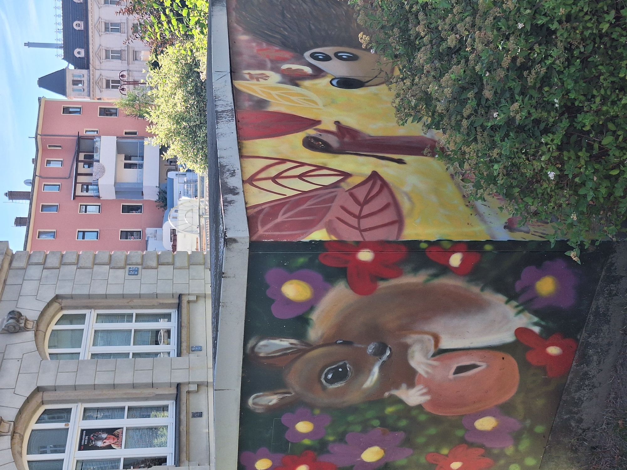 The image shows a street art mural on a concrete wall. The mural features cartoonish depictions of animals and figures. On the left side, a brown squirrel is holding an orange object, surrounded by purple and red flowers. To the right, a figure resembling a hedgehog with large eyes is depicted, along with a small human-like figure in a red dress and stylized leaves in shades of red and yellow. The mural is located in the city of Aue, Germany, as indicated by context. In the background, there are buildings, including a pink multi-story building and a cream-colored building with a dark roof and spire. The sky is blue and clear. Green bushes are growing in front of the mural.
